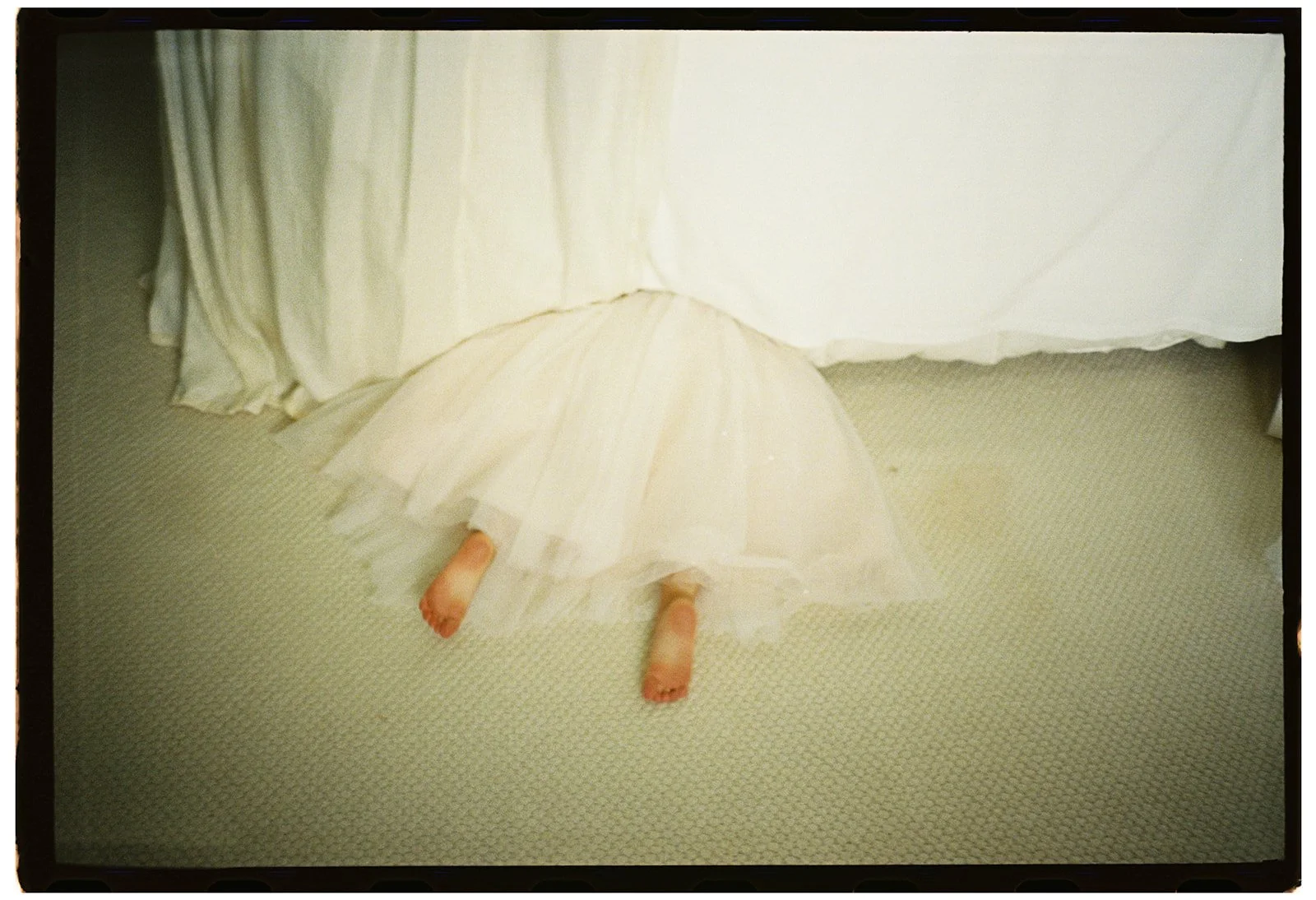 A flower girl hiding beneath a wedding dress with only her tiny feet visible, capturing a playful and candid moment during wedding preparations.