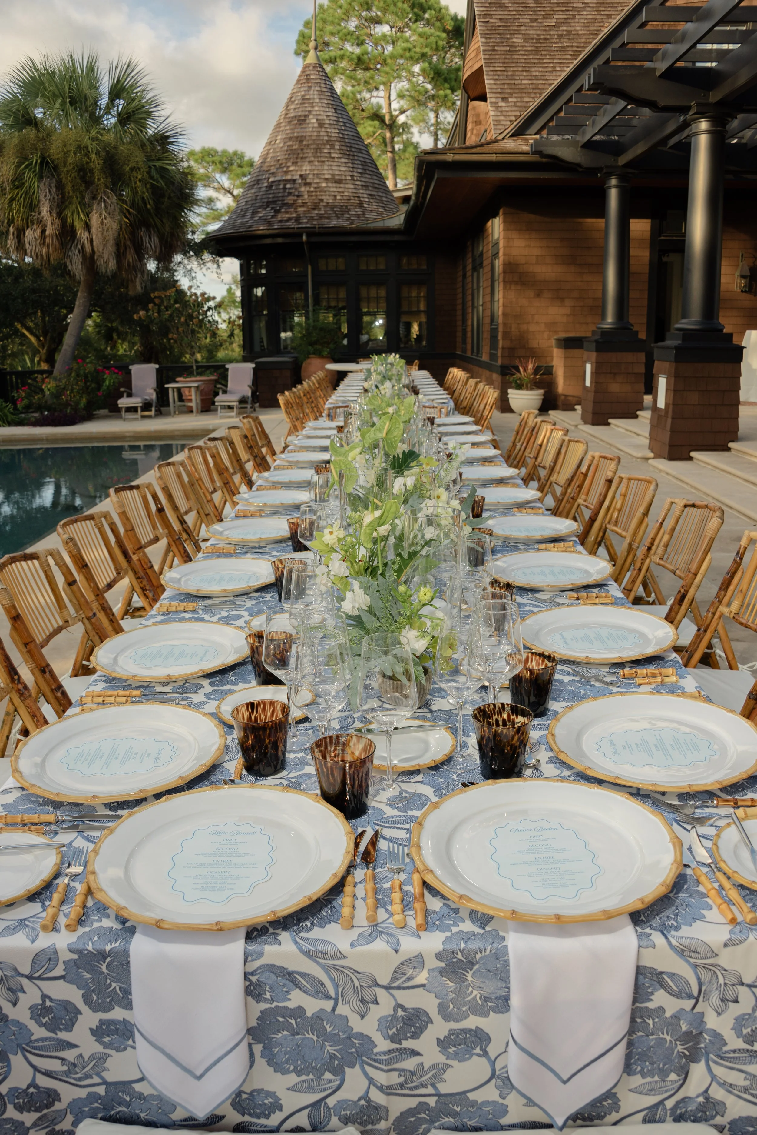 Long rehearsal-dinner table featuring blue botanical linens, scalloped menus, gold-rimmed plates, taper candles, and floral designs by Festoon.