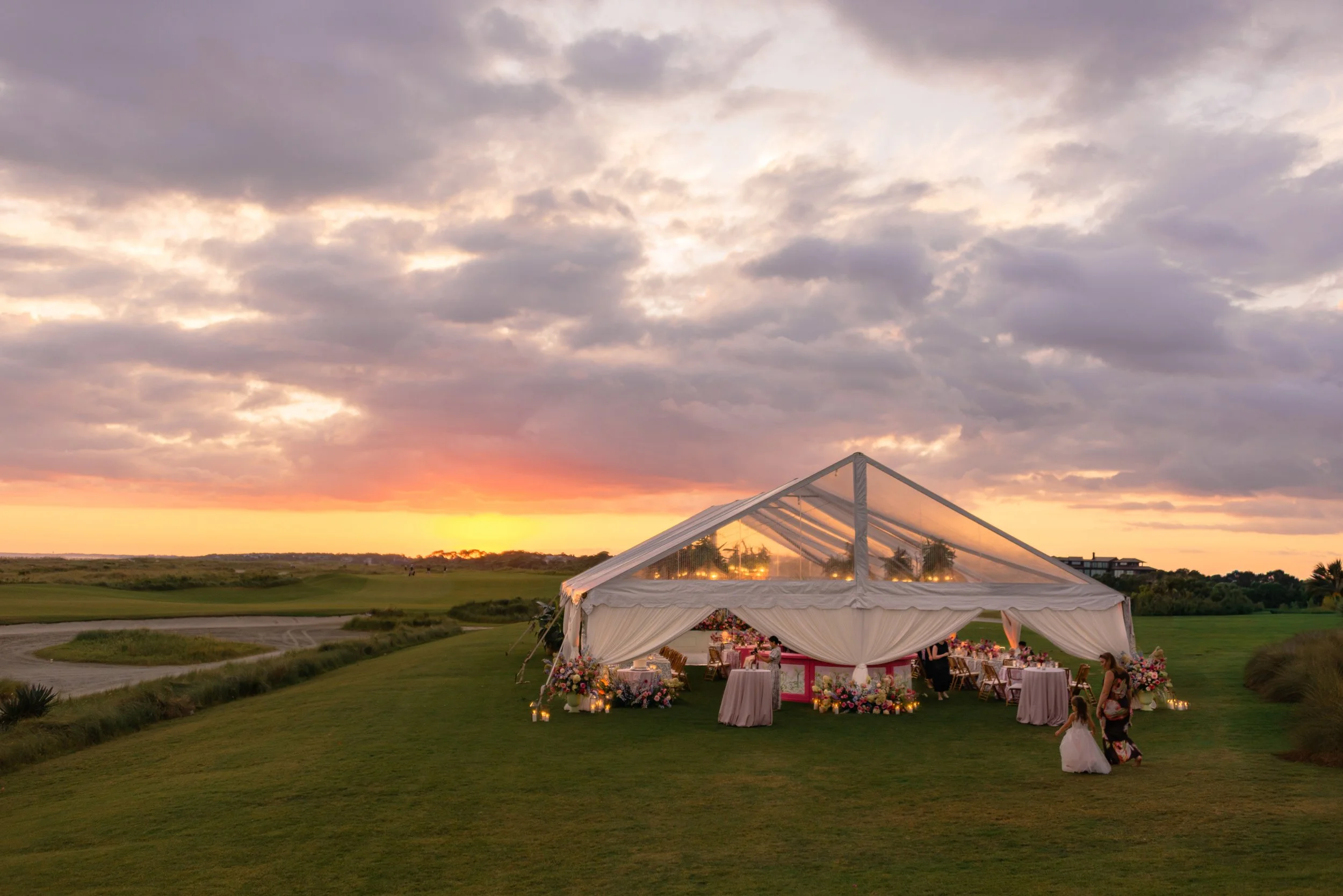 Clear-top wedding reception tent glows beneath a dramatic pink-and-orange sunset at The Ocean Course on Kiawah Island.