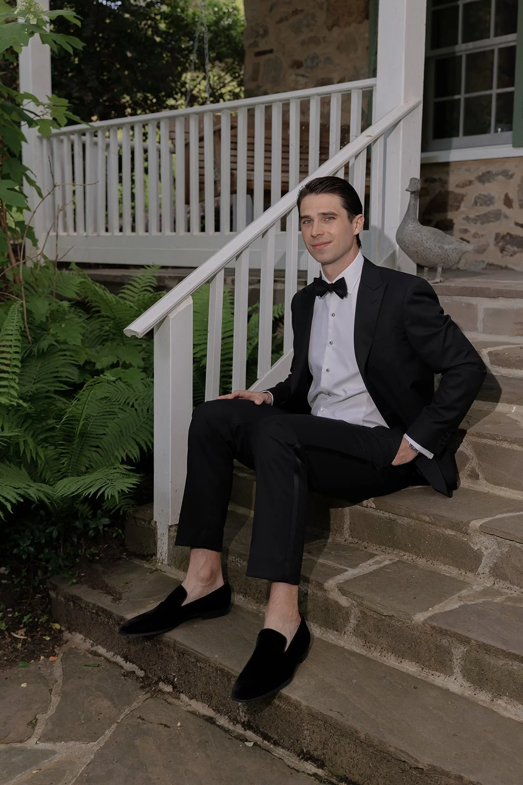 Groom seated on a stone step outdoors at the Spring House at Goodstone Inn in a black tuxedo.