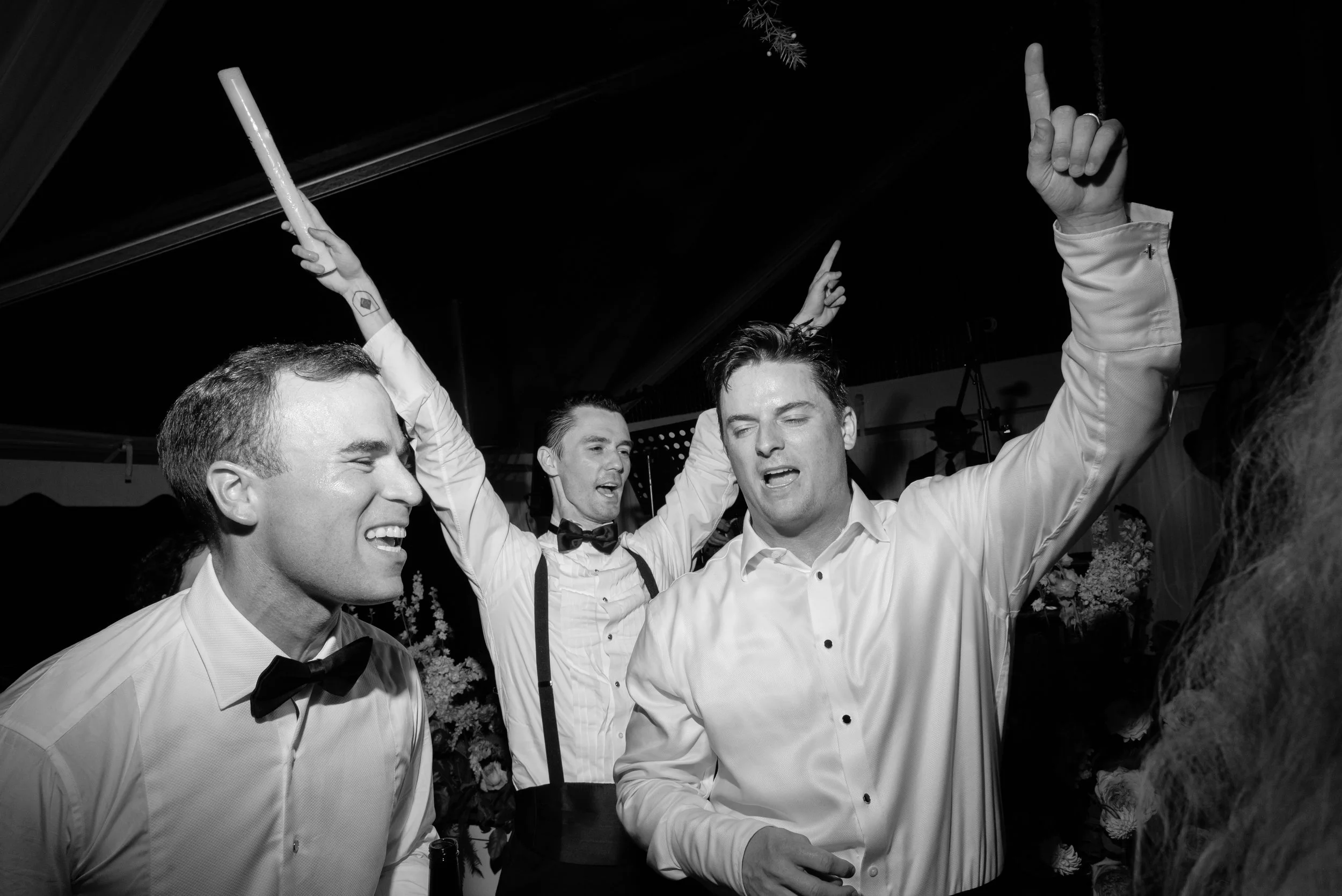 Black-and-white photograph of wedding guests singing and raising their arms on the crowded dance floor.