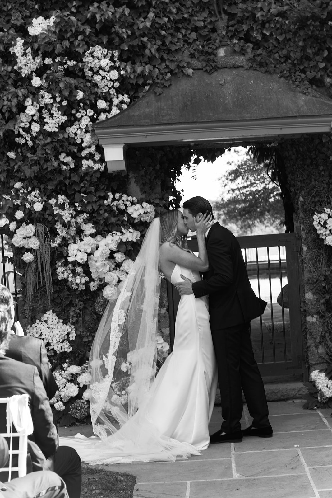 Black-and-white photograph of the bride and groom sharing their first kiss beneath the floral Ivy Wall ceremony setting at Goodstone Inn.