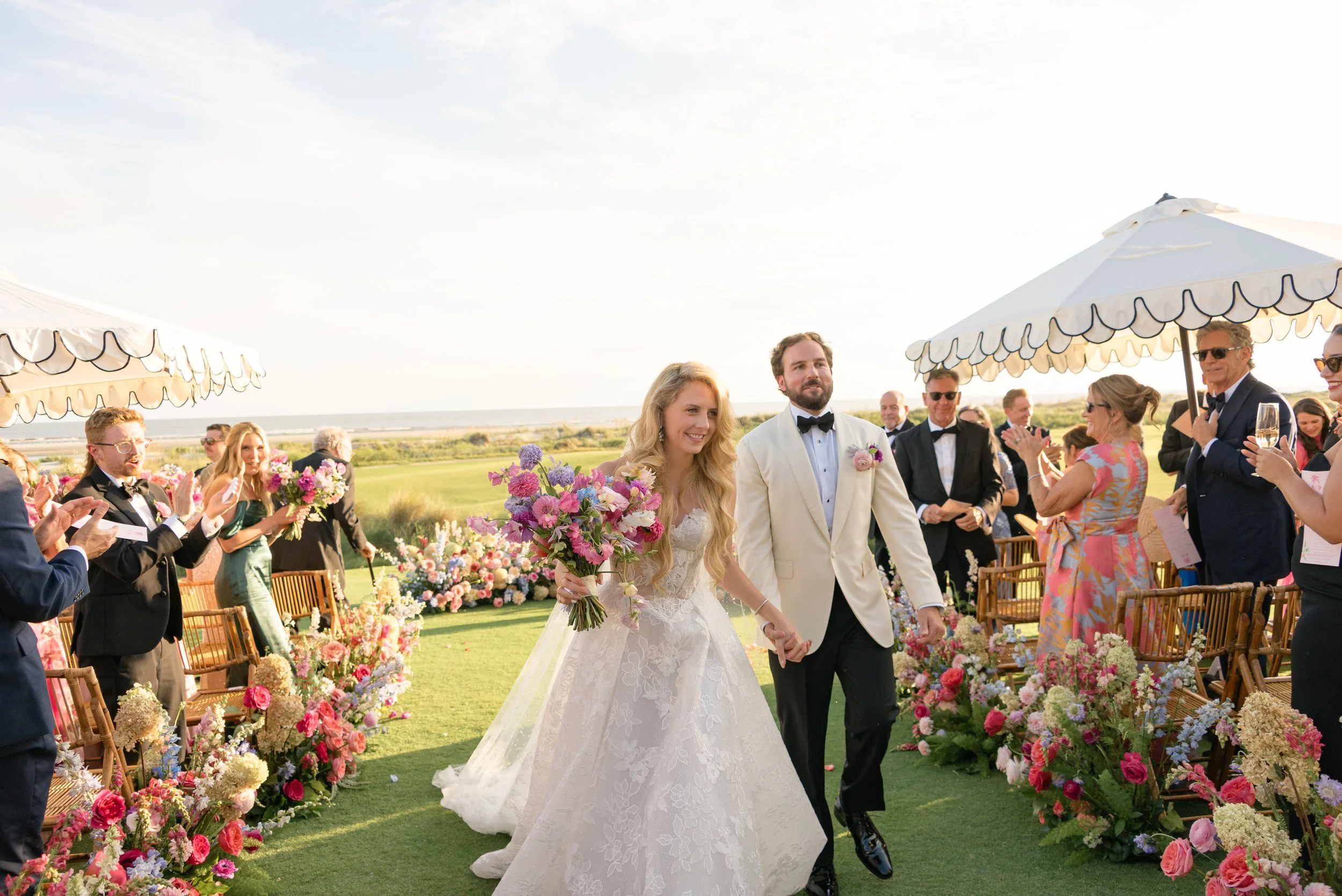 Newlyweds walk through a colorful floral aisle after their wedding ceremony at The Ocean Course on Kiawah Island, with flowers by Festoon.