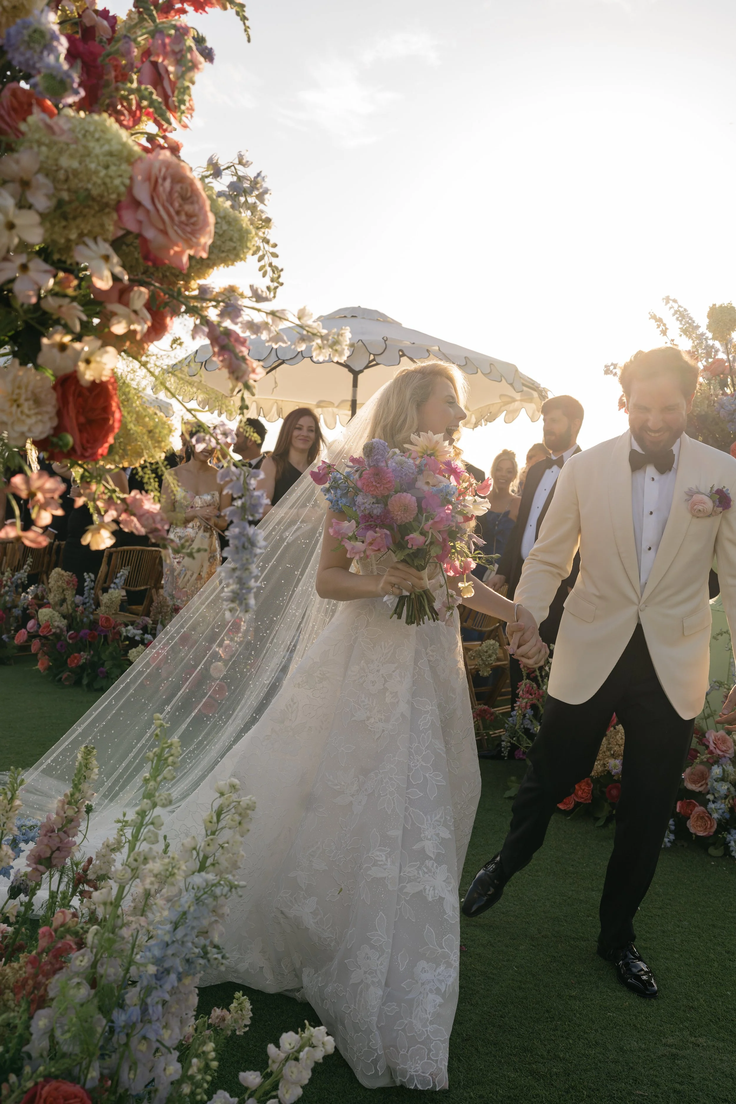 Newlywed couple walking together through a colorful floral wedding installation during their destination wedding celebration.