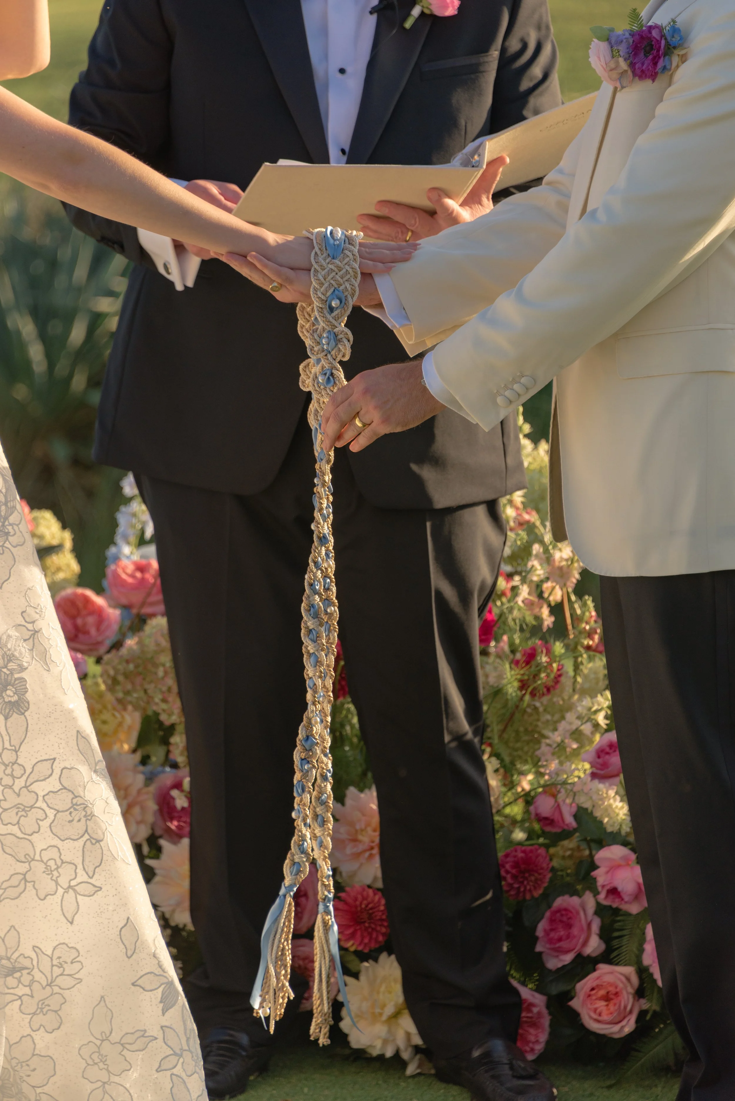 Close-up view of Katie and Trevor’s hand-fasting during their wedding ceremony, framed by colorful flowers at The Ocean Course.