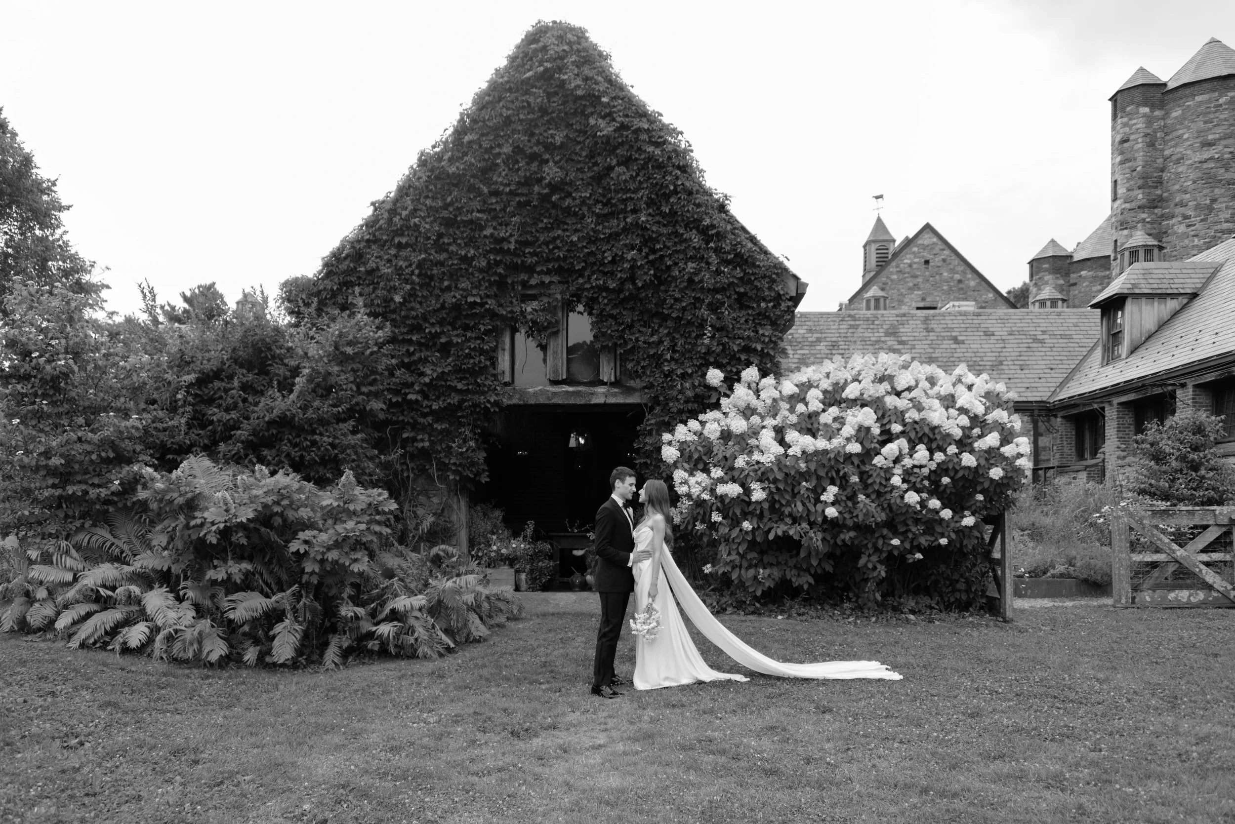 An editorial black-and-white, timeless photo of the bride and groom and her dramatic cape outside the iconic design of Blue Hill at Stone Barns with the white summer hydrangeas blooming in the background. 