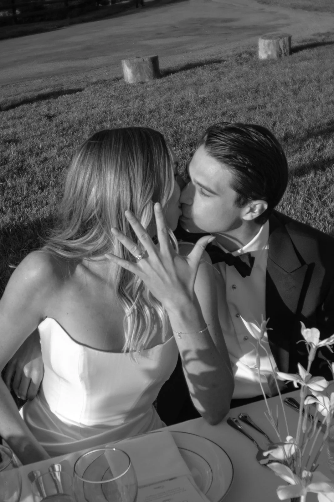 Black-and-white photo of the bride and groom kissing during dinner why the bride shows her hand with her new wedding band on it to the camera