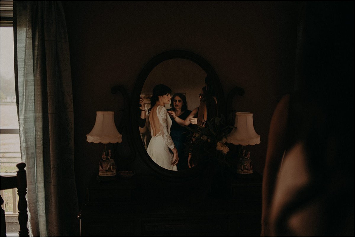  The bride’s reflection in the mirror as her bridesmaids help fasten her gown 
