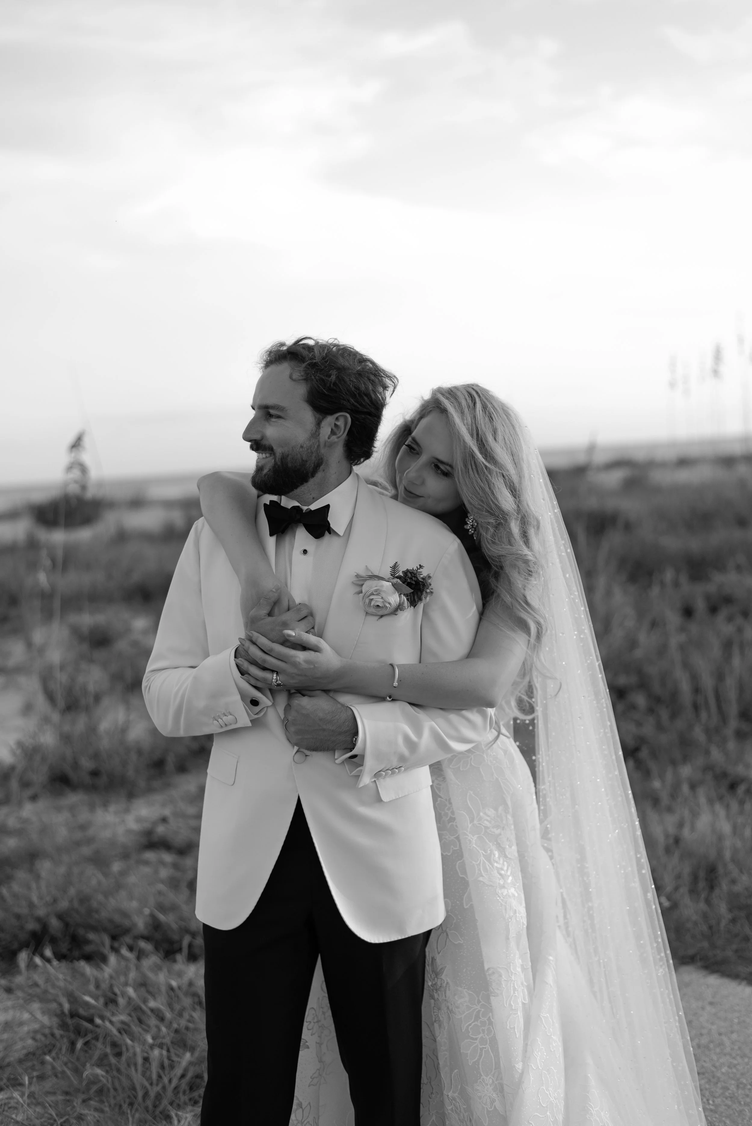 Black-and-white portrait of Katie and Trevor embracing against the coastal grasslands at The Ocean Course.