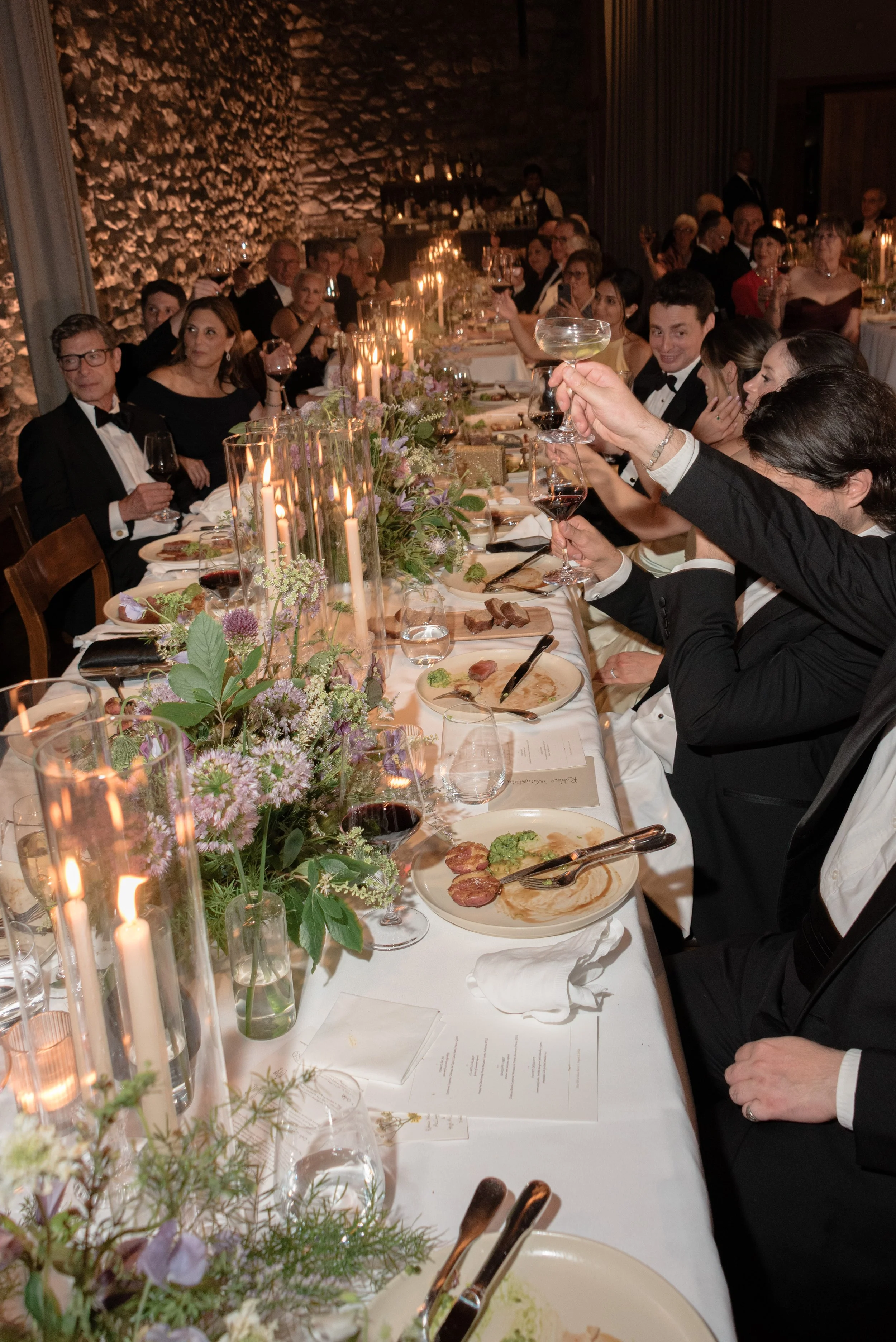 An editorial flash photo of the long reception head table filled with flowers, candles, wine and guests toasting during dinner at Blue Hill at Stone Barns.