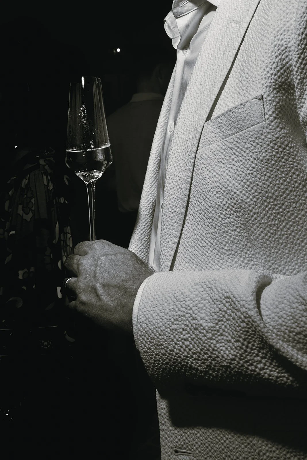 Black and white film photograph of a groom in a textured white tuxedo jacket holding a champagne glass during a Charleston wedding celebration, capturing an intimate editorial moment.