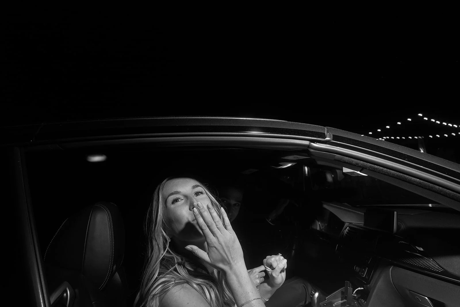 Black-and-white portrait of the bride seated inside a getaway car and blowing a kiss to her wedding guests at the end of her Goodstone Inn wedding night.