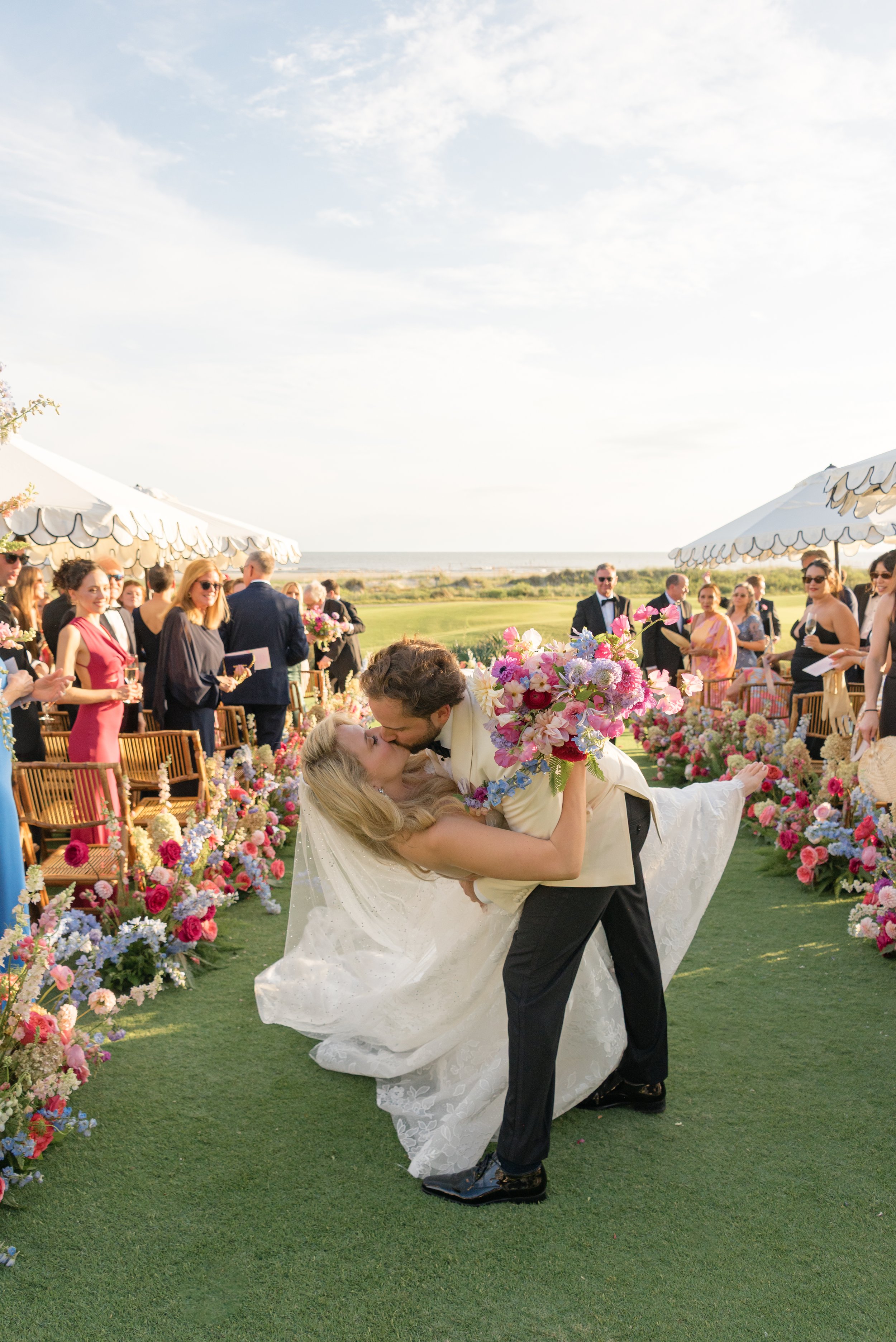 Trevor dips Katie for a celebratory kiss in the floral aisle as their guests applaud after the Kiawah Island ceremony.