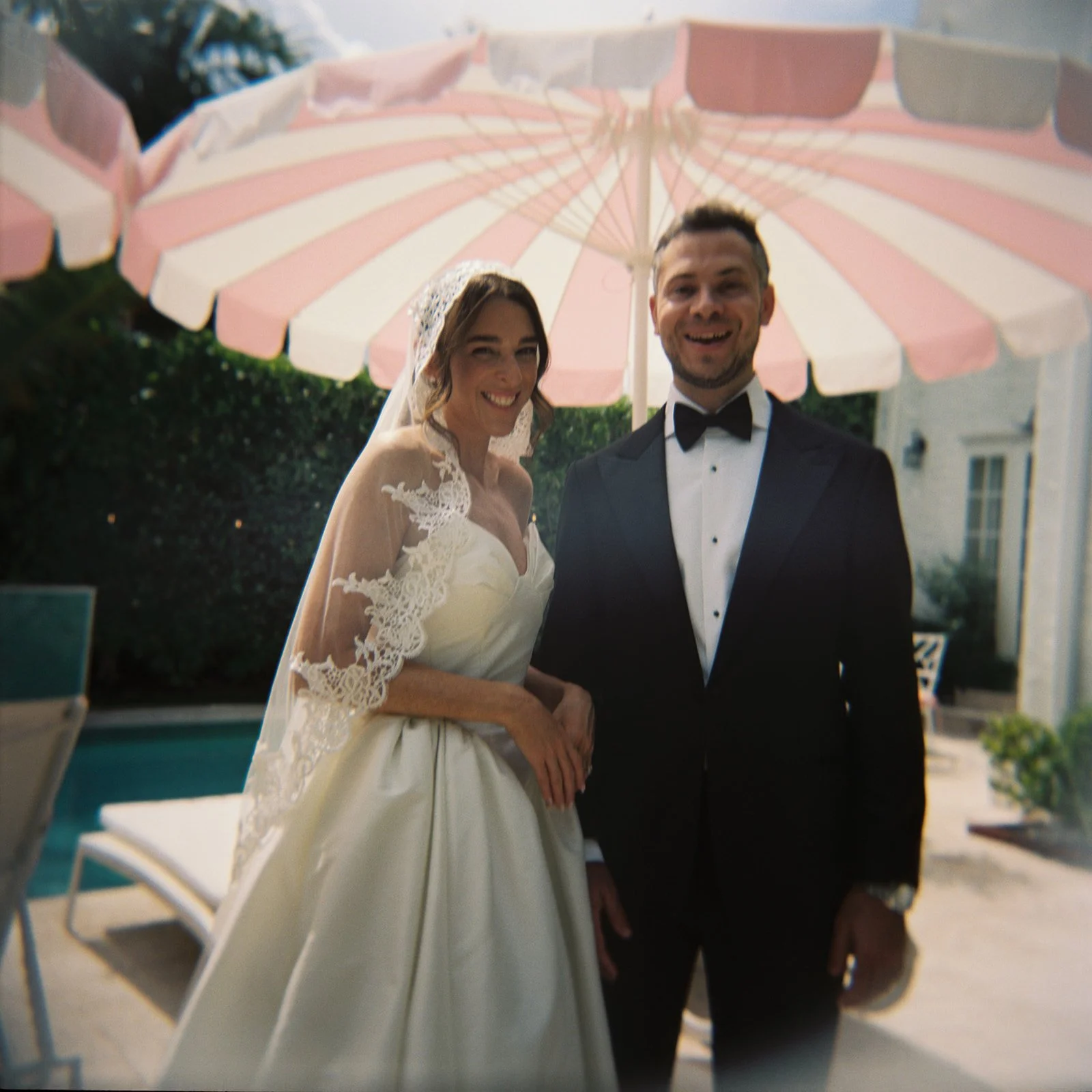 Holga film photograph of a newlywed couple smiling together beneath a pink and white striped umbrella during their Orthodox Jewish wedding celebration in West Palm Beach, Florida, following their reception at Malka restaurant.