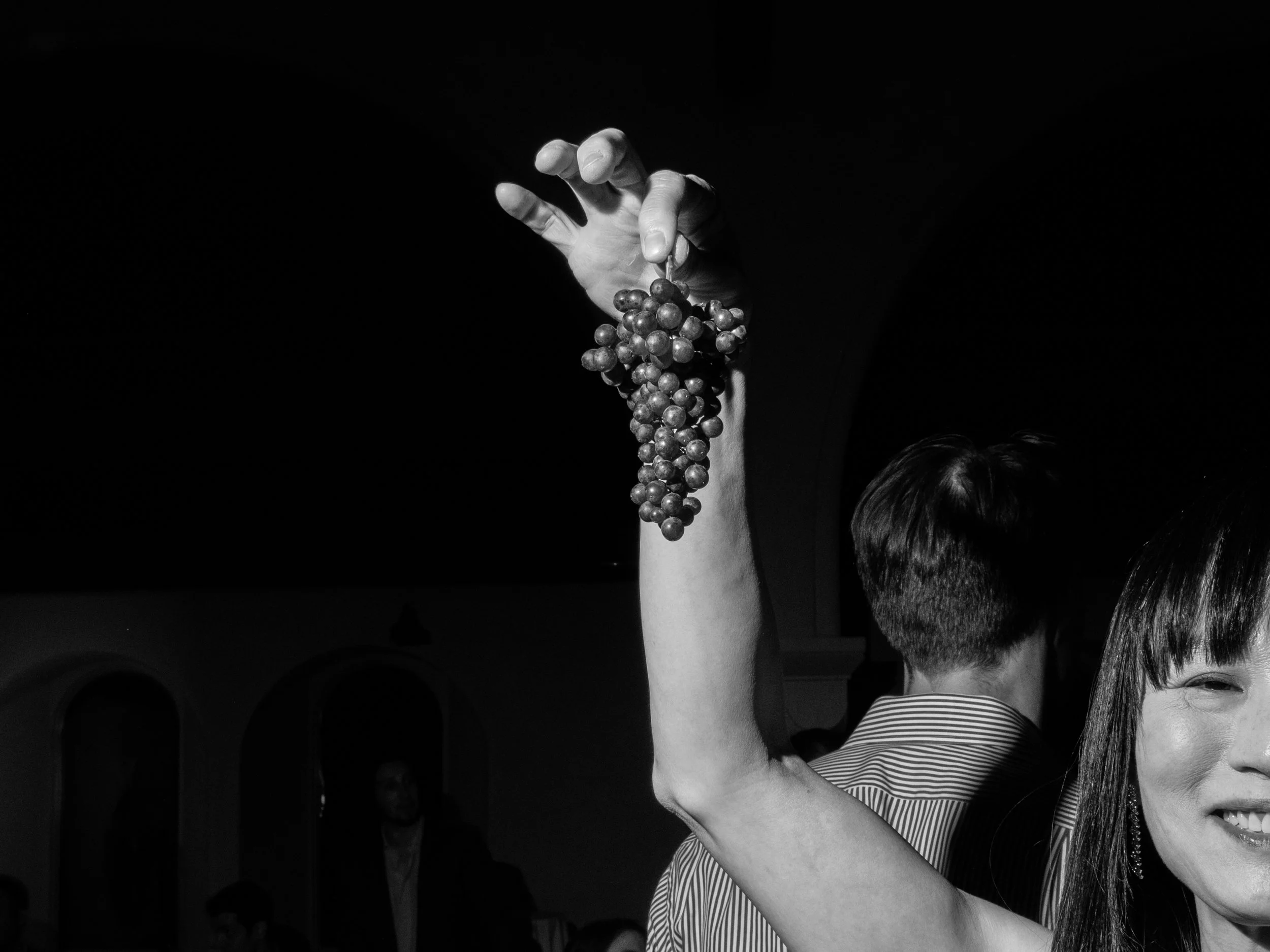 A black and white editorial photograph of a woman holding a bunch of grapes from the food display table.