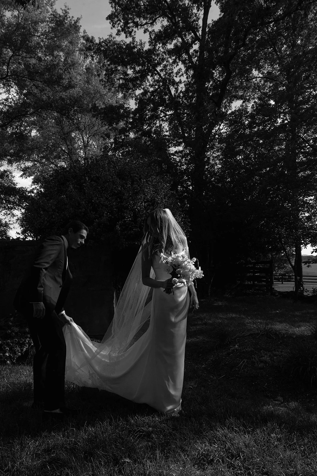 Black-and-white portrait of the groom adjusting the bride's wedding veil and dress train.