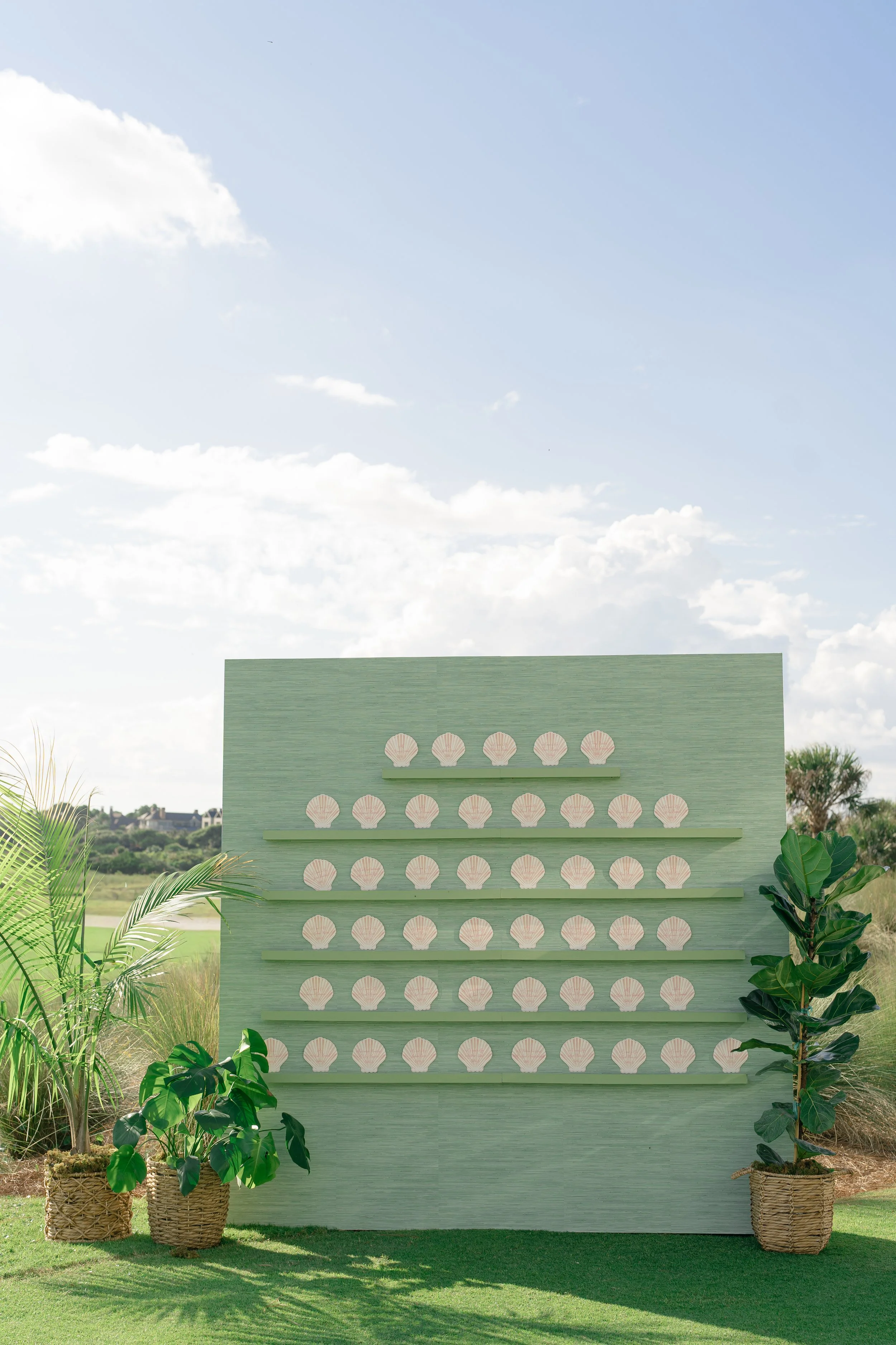 Pale green escort-card wall decorated with rows of white seashells and tropical plants for a coastal Kiawah Island wedding.