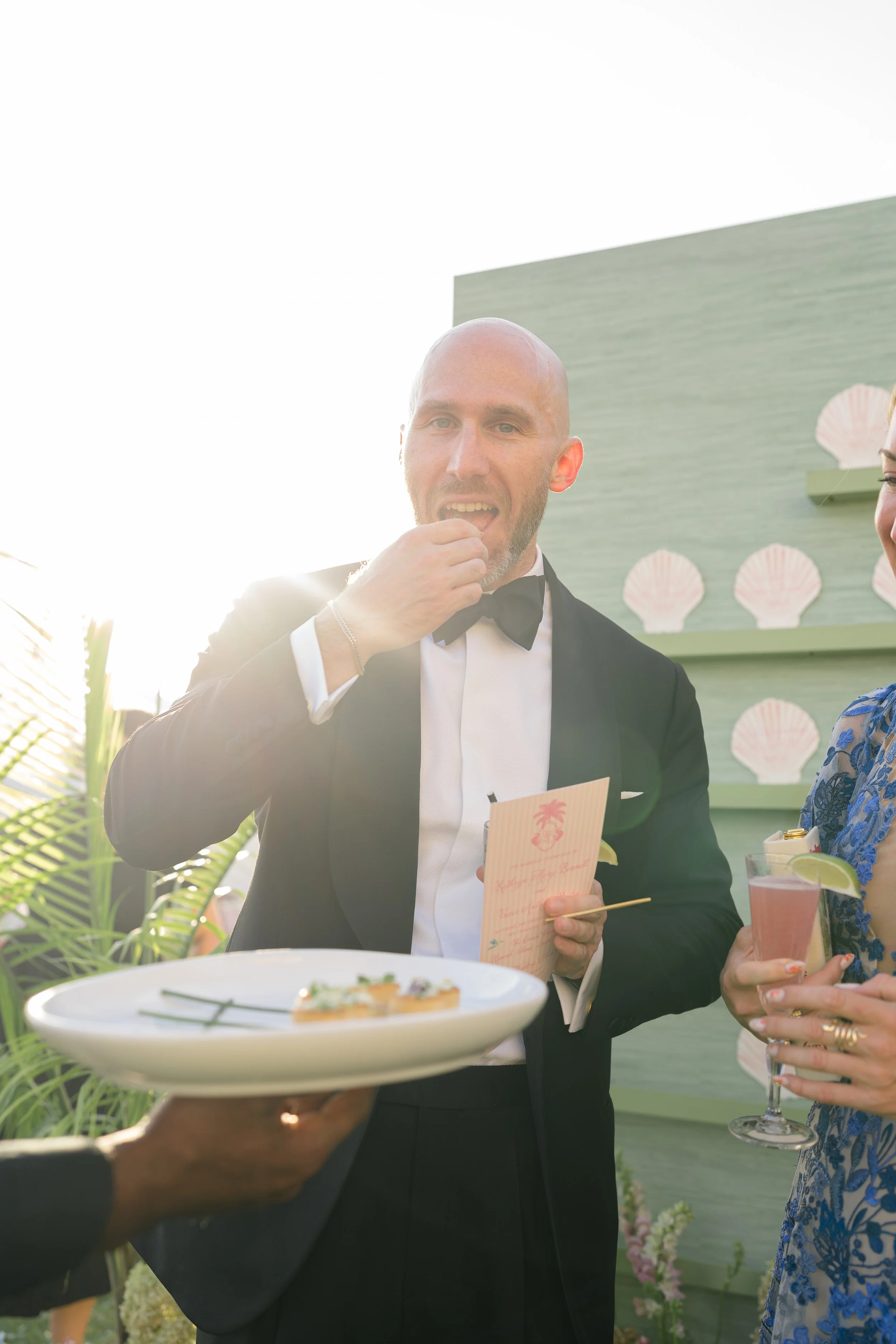 Black-tie wedding guest enjoys a passed hors d’oeuvre beside the seashell escort display during cocktail hour.