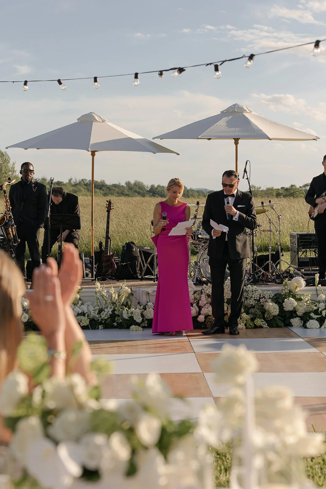 The parents of the bride give a speech during the reception dinner beneath two ivory umbrellas and string lights on a checkerboard dance floor in the Hayfield at Goodstone Inn.