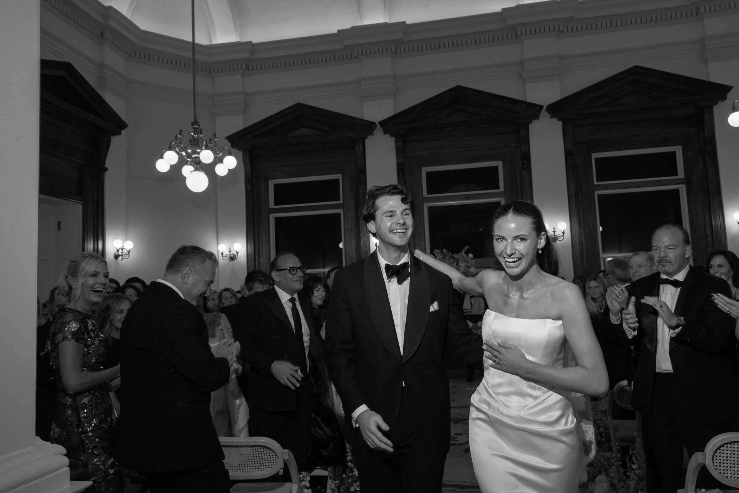 Black-and-white direct-flash photograph of newlyweds exit their wedding ceremony as guests celebrate around them at the Gibbes Museum in Charleston, SC.