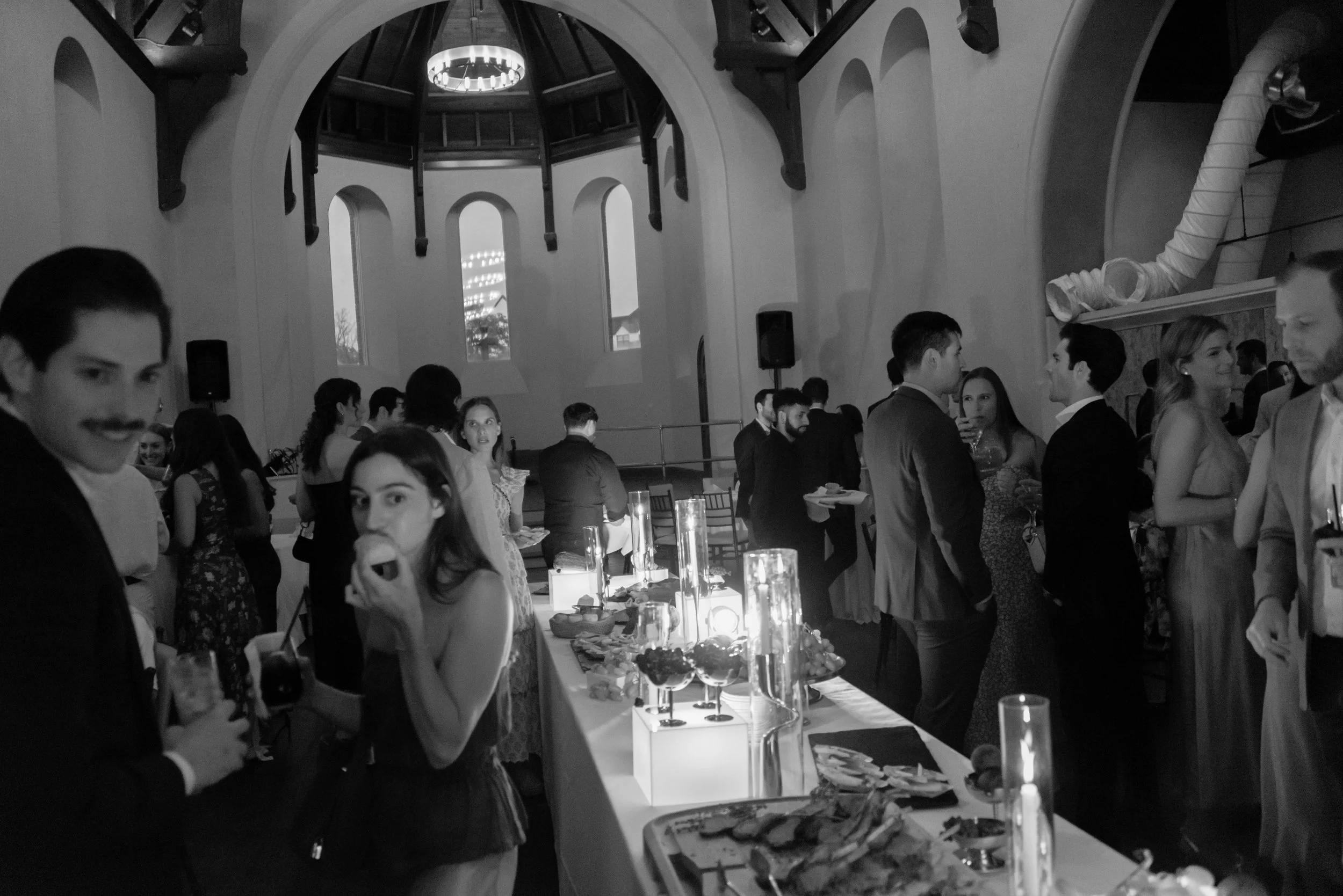 Black-and-white documentary photograph of guests mingling around the long dinner table.