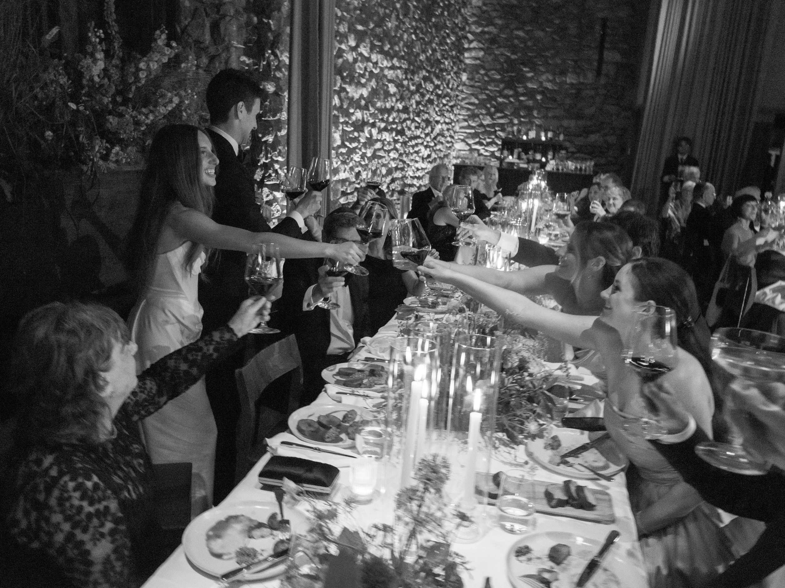 Black-and-white overhead view of guests toasting wine glasses around the floral-filled reception table.
