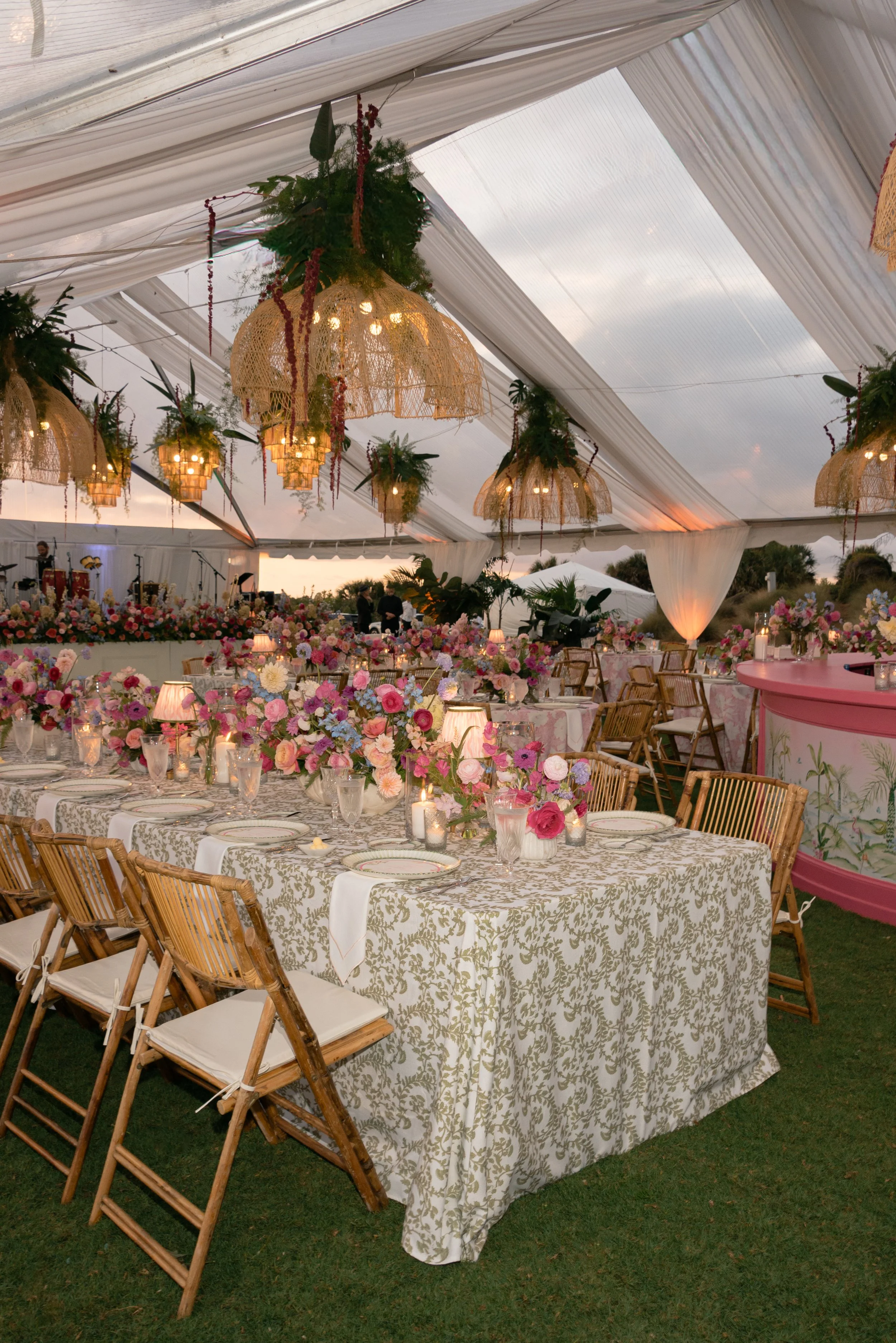 Clear-top reception tent filled with patterned linens, rattan chairs, woven chandeliers, and abundant flowers by Festoon.