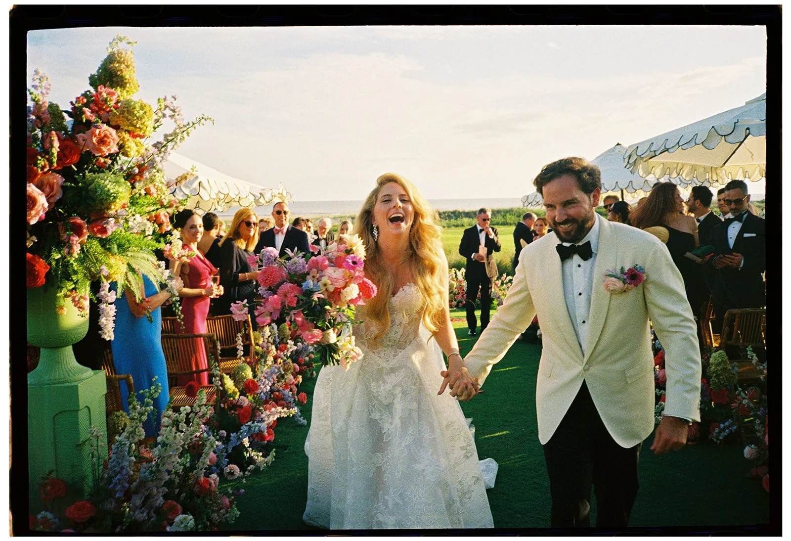 35mm film photograph of a bride and groom walking together after their wedding ceremony at The Ocean Course on Kiawah Island, South Carolina, surrounded by colorful floral designs and guests during their multi-day destination wedding celebration.