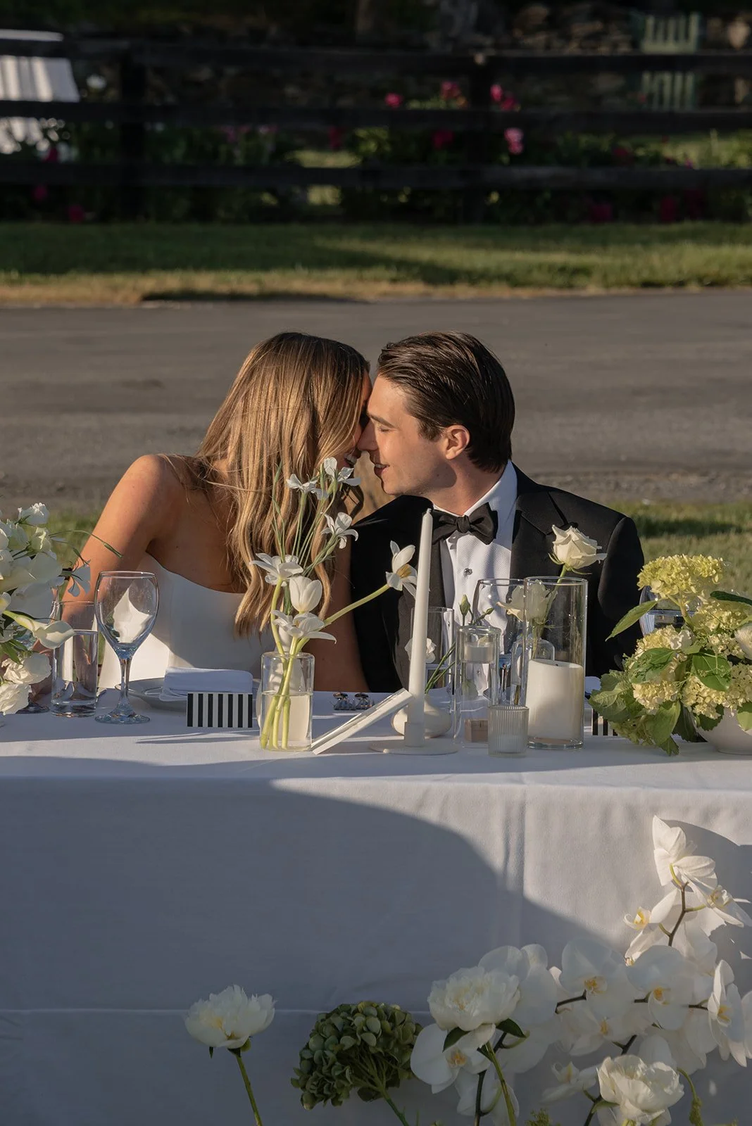 Bride and groom share a kiss during the speeches at an outdoor reception table surrounded by white flowers and glassware at Goodstone Inn.