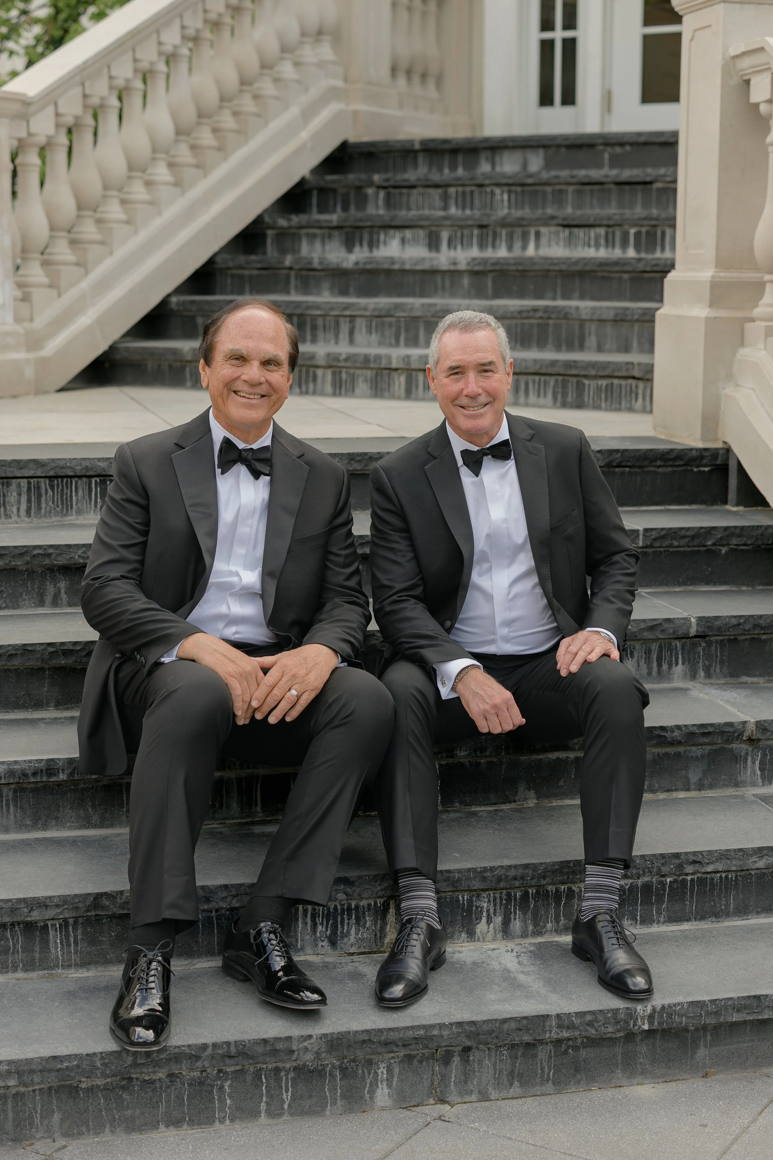 The father of the bride and the father of the groom in classic black-tie attire seated on the steps at Shadowbrook at Shrewsbury.