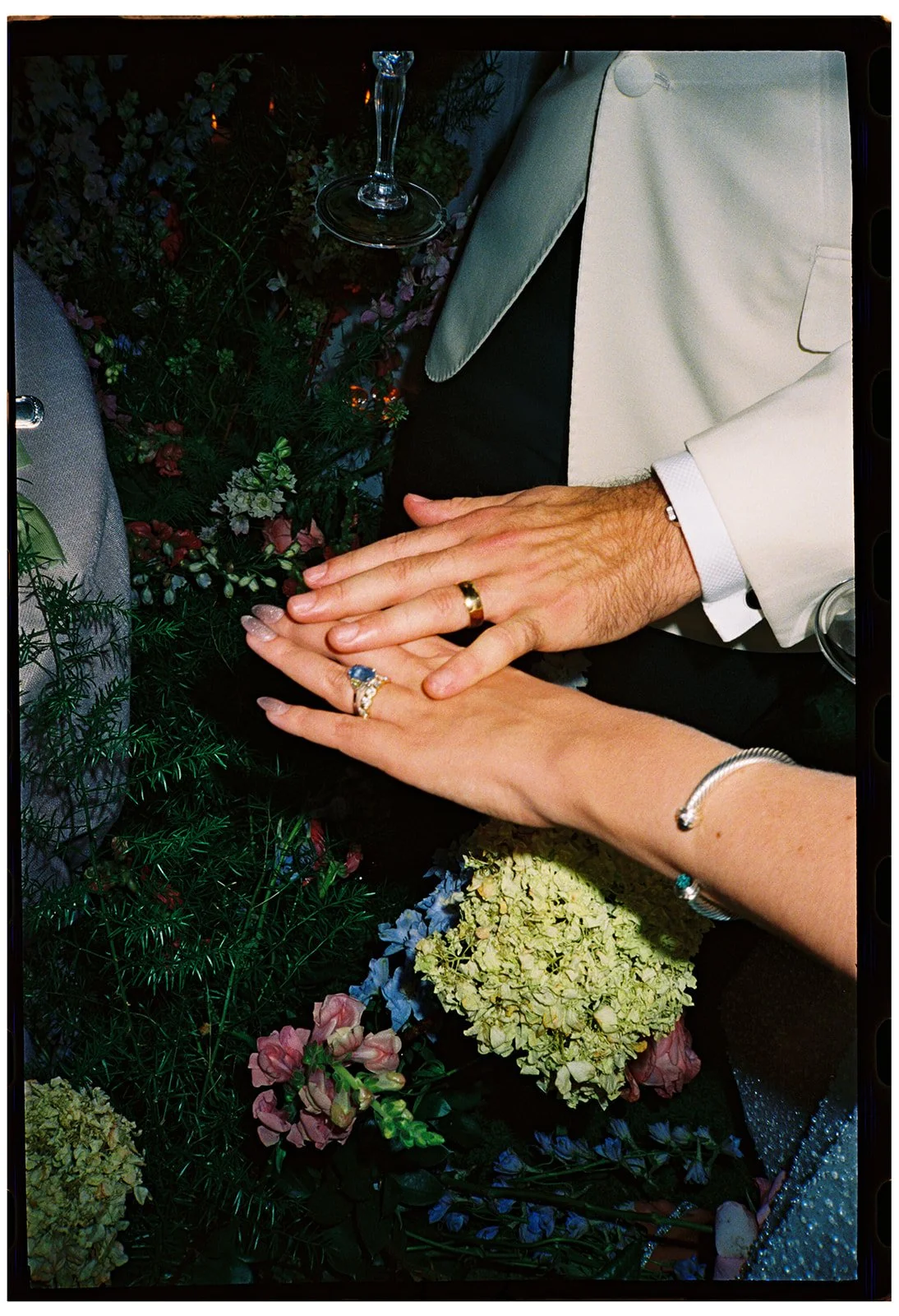 Close-up editorial photograph of newlywed hands and wedding rings surrounded by floral details, capturing the intimate textures of a celebration.