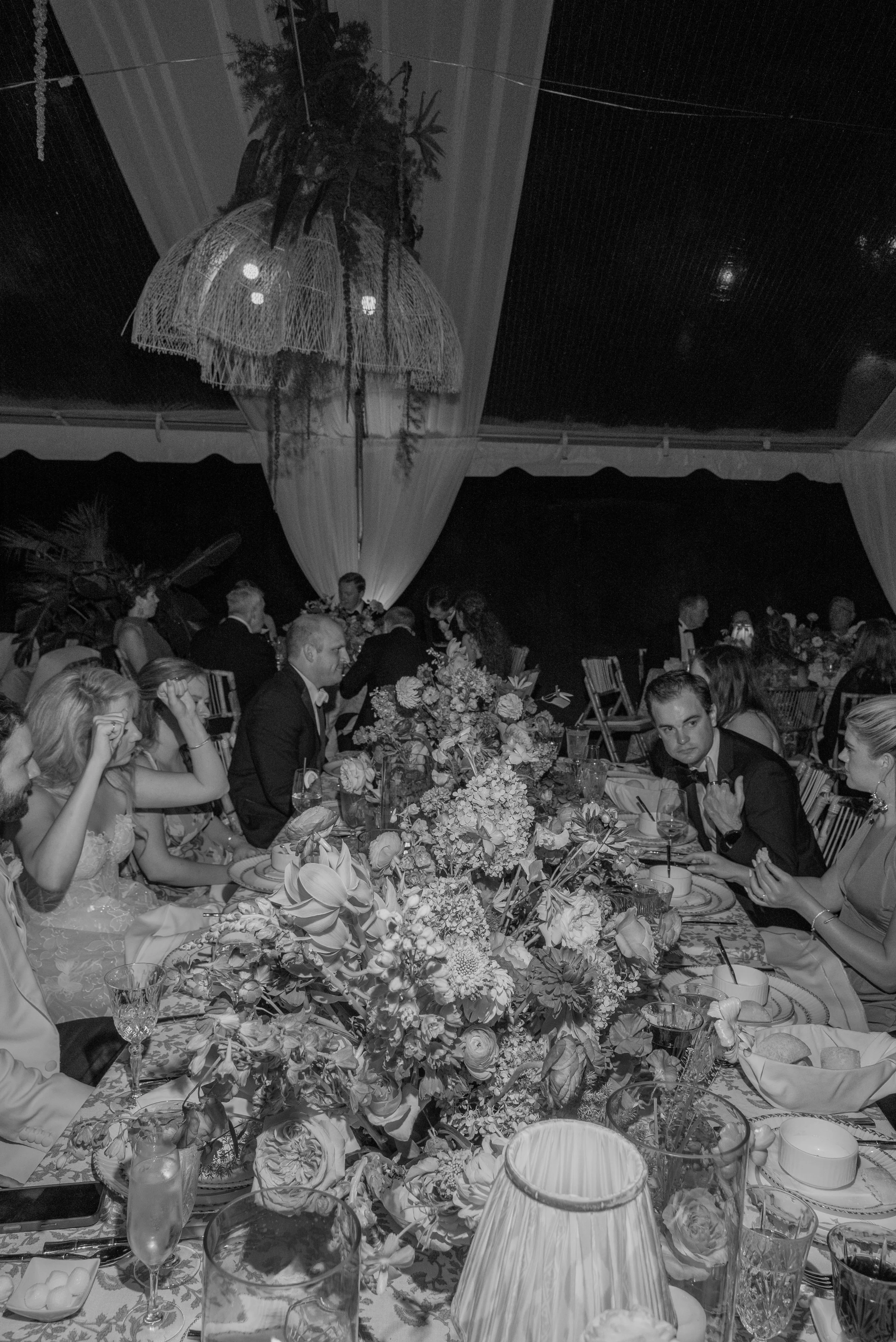 Black-and-white photograph of guests gathered around a richly layered floral table during dinner.