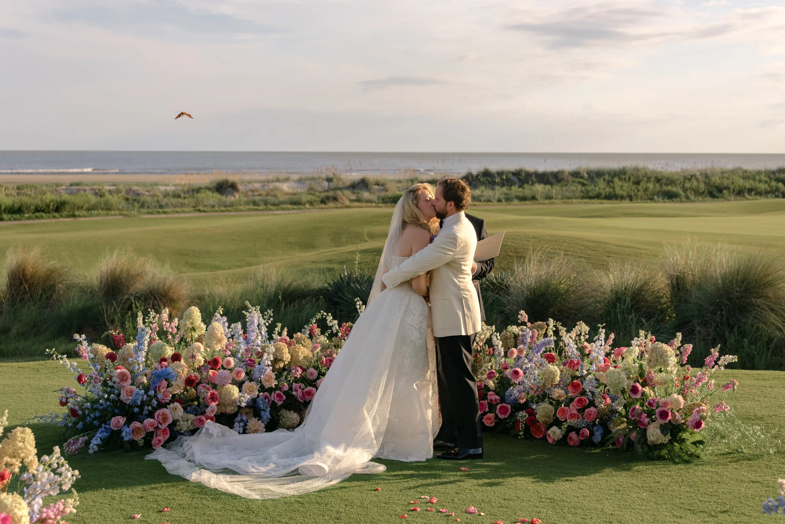 The bride and groom kiss inside a sweeping semicircle of colorful flowers by Festoon with the Kiawah Island landscape behind them and a perfectly timed butterfly photo-bomb.