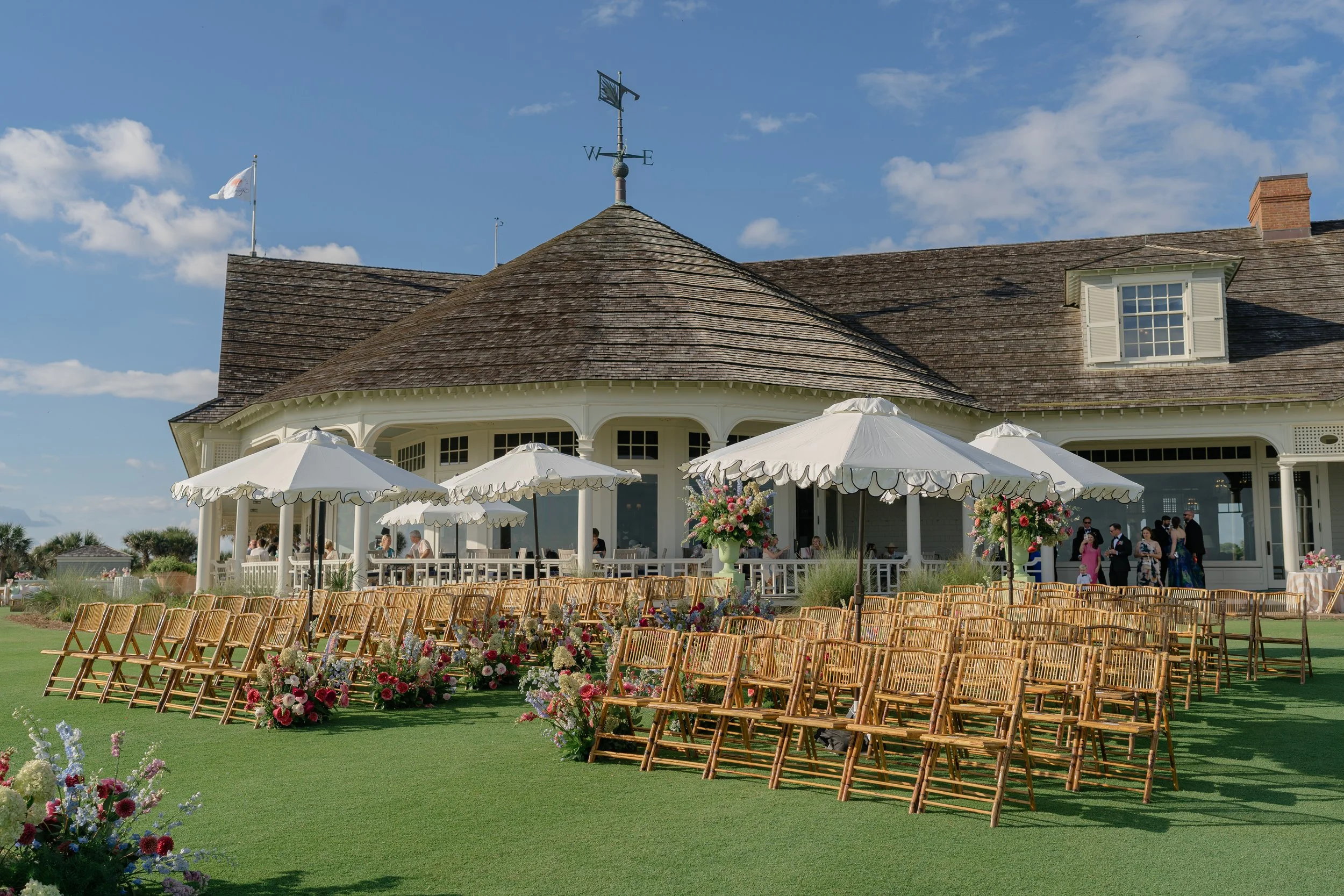 Ocean Course clubhouse framed behind the outdoor ceremony lawn, scalloped umbrellas, wooden chairs, and colorful floral aisle.