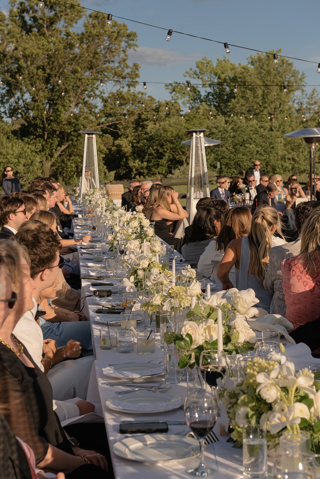 Wedding guests seated along a long outdoor dinner table with white flowers and glassware at Goodstone Inn.