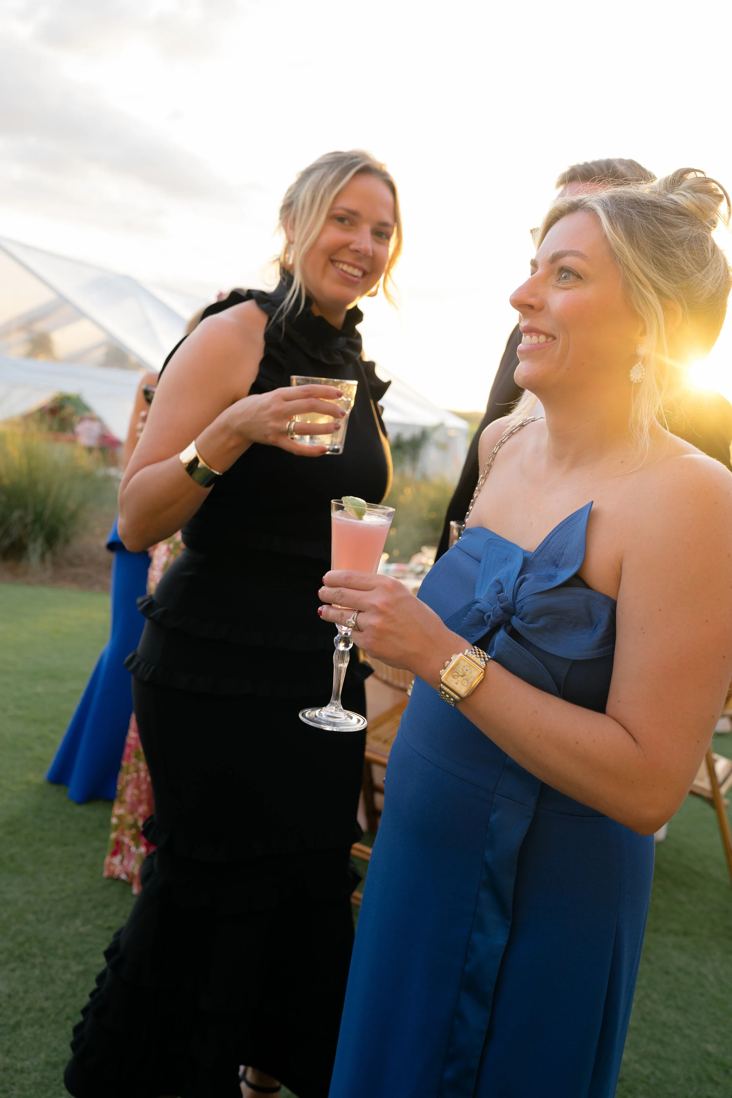 Two wedding guests in evening dresses share cocktails and conversation in the golden light.