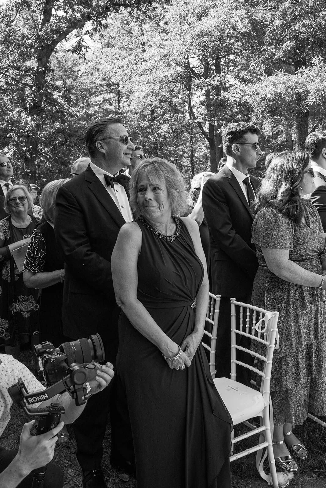 Black-and-white candid of groom's mother looking back at him while he watches the bride walk down the aisle.