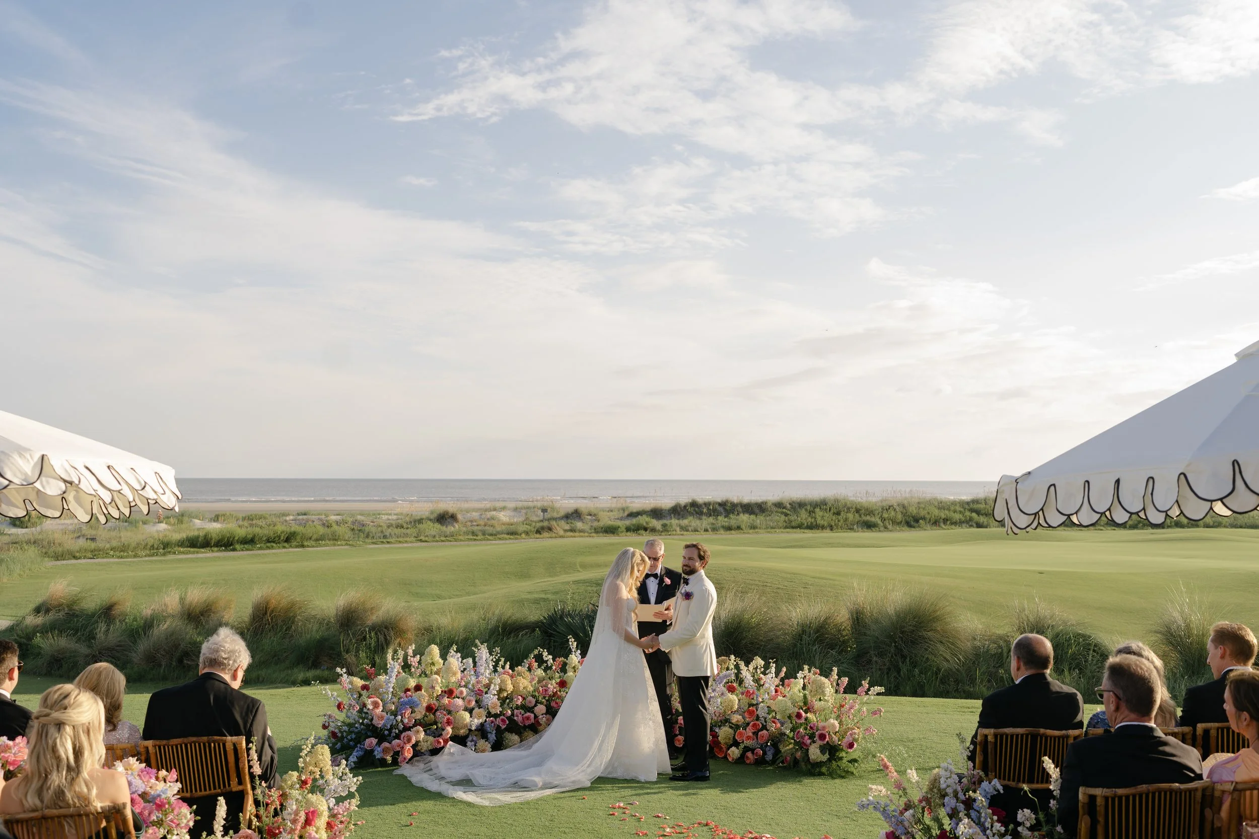 The bride and groom stand together within a semicircle of colorful flowers during their oceanfront wedding ceremony on Kiawah Island.