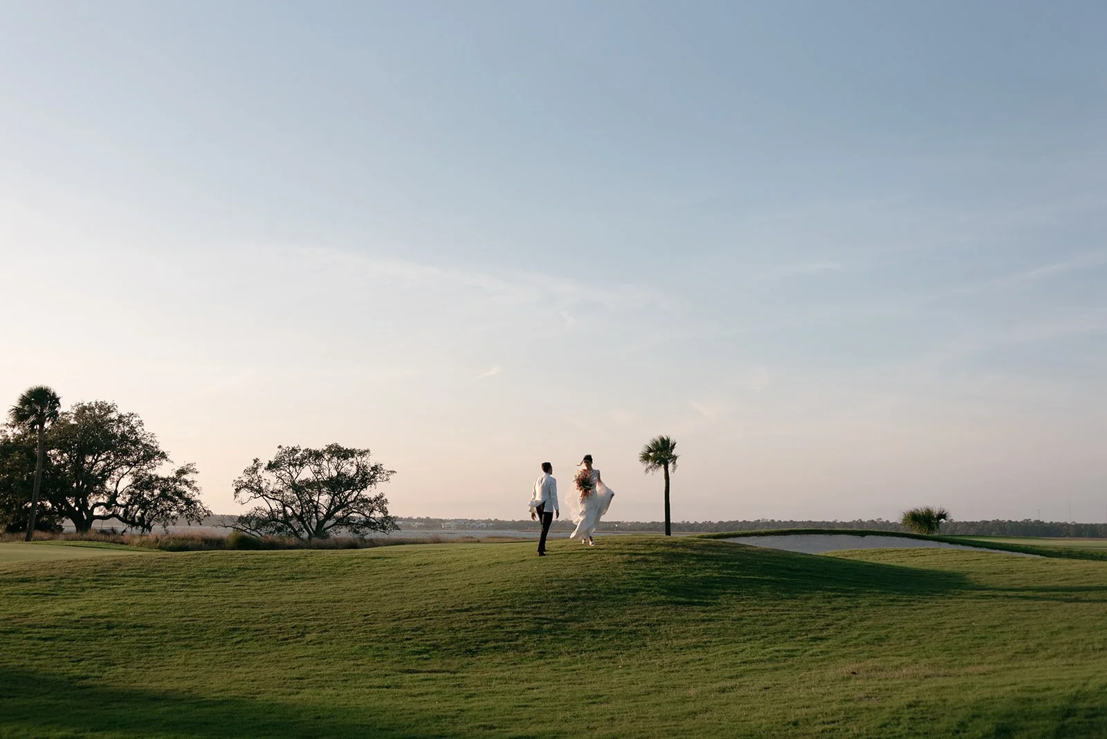 A newlywed couple walking together across a grassy landscape at sunset after their wedding on Kiawah Island, South Carolina.