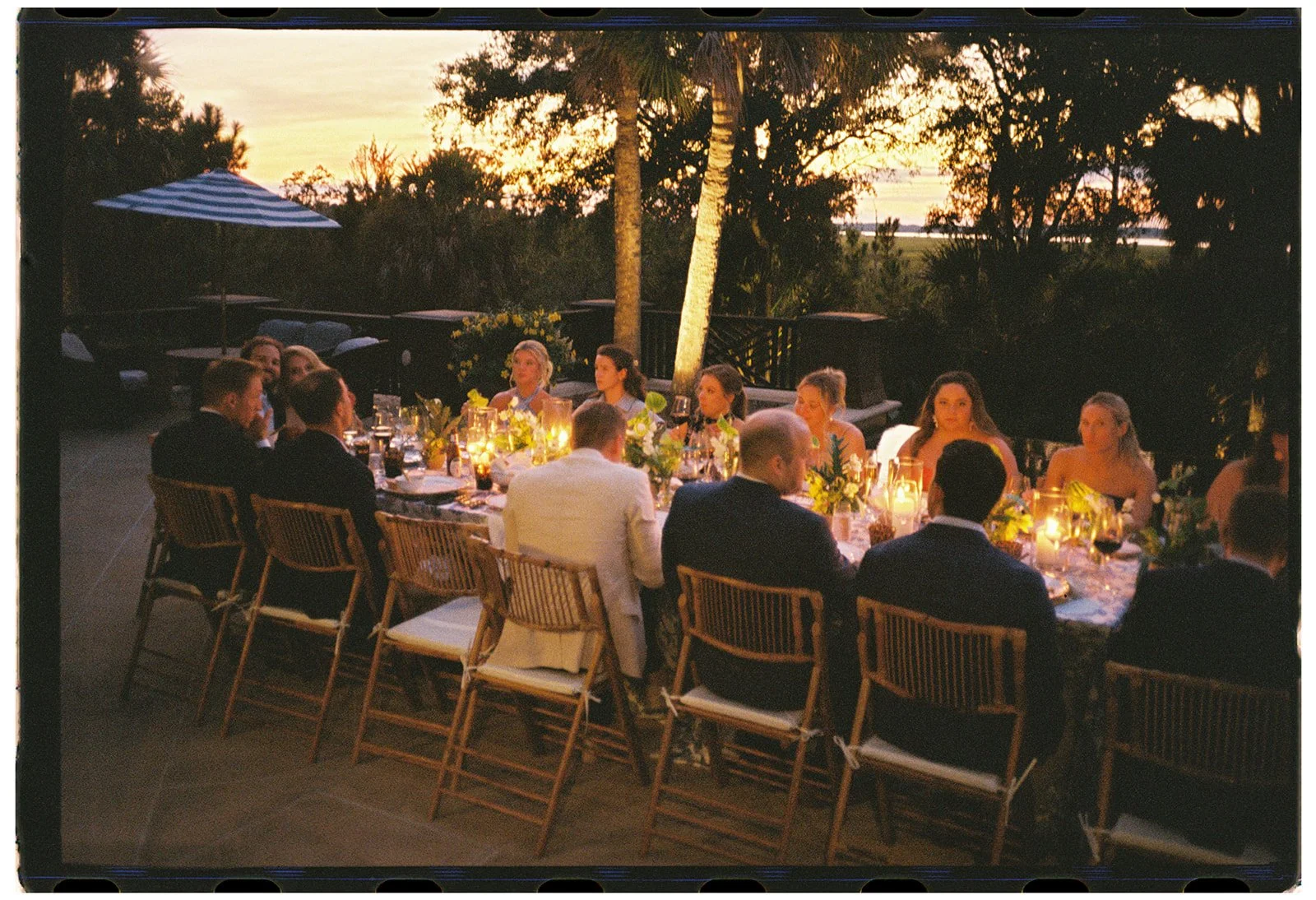 Outdoor rehearsal dinner beneath palm trees and evening light, with guests gathered around a candlelit table at a private estate on Kiawah Island in a destination celebration setting.