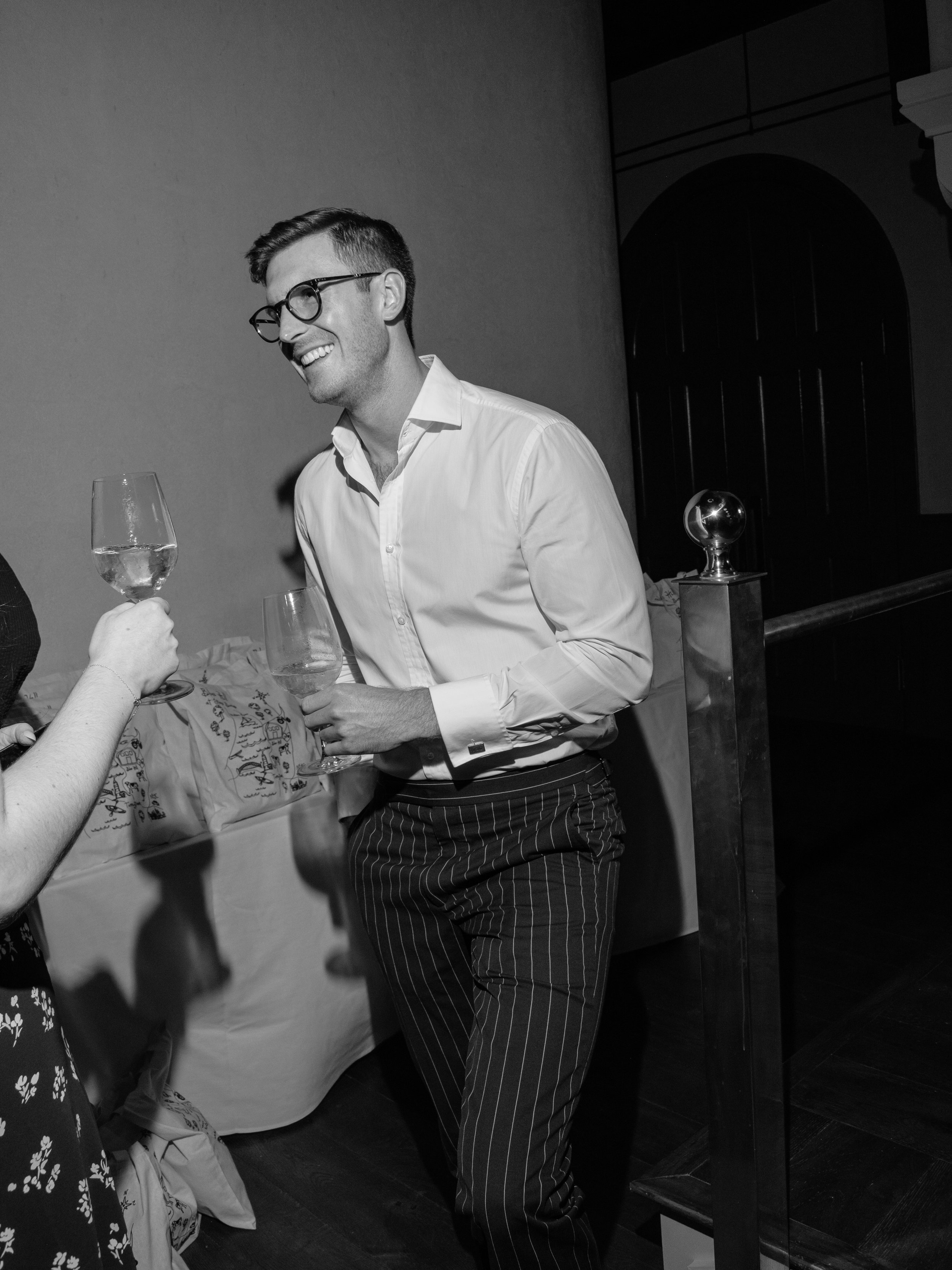 Fashionable male guest in pinstripe navy pants and a white button down with a wine glass in hand laughs with another guest during the rehearsal dinner in Hudson Valley.