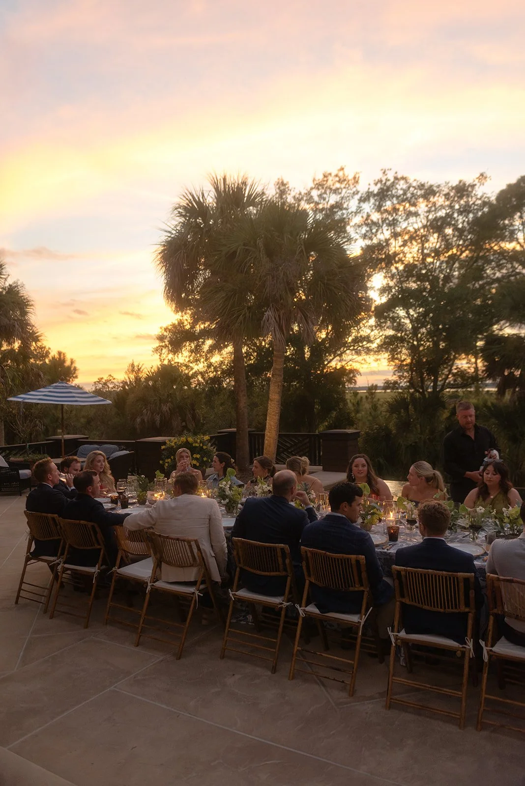 Wedding guests gather around a long outdoor dinner table beneath palm trees during a glowing sunset rehearsal dinner.