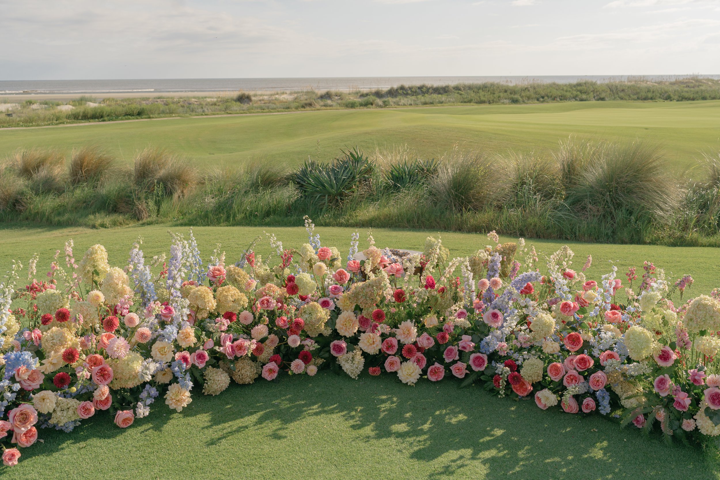 Dense arrangement of pink, lavender, cream, and coral flowers by Festoon borders the Ocean Course wedding ceremony.