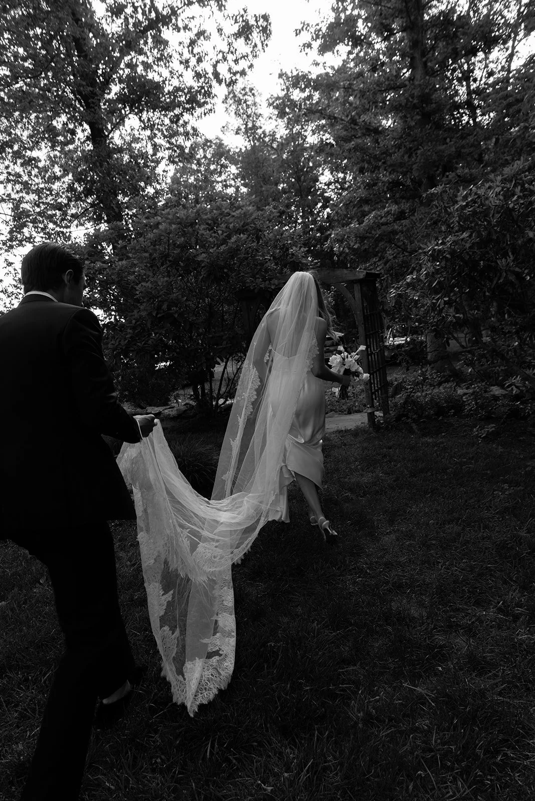 Black-and-white photograph of the bride walking through the gardens at Goodstone Inn with her long veil trailing behind her.