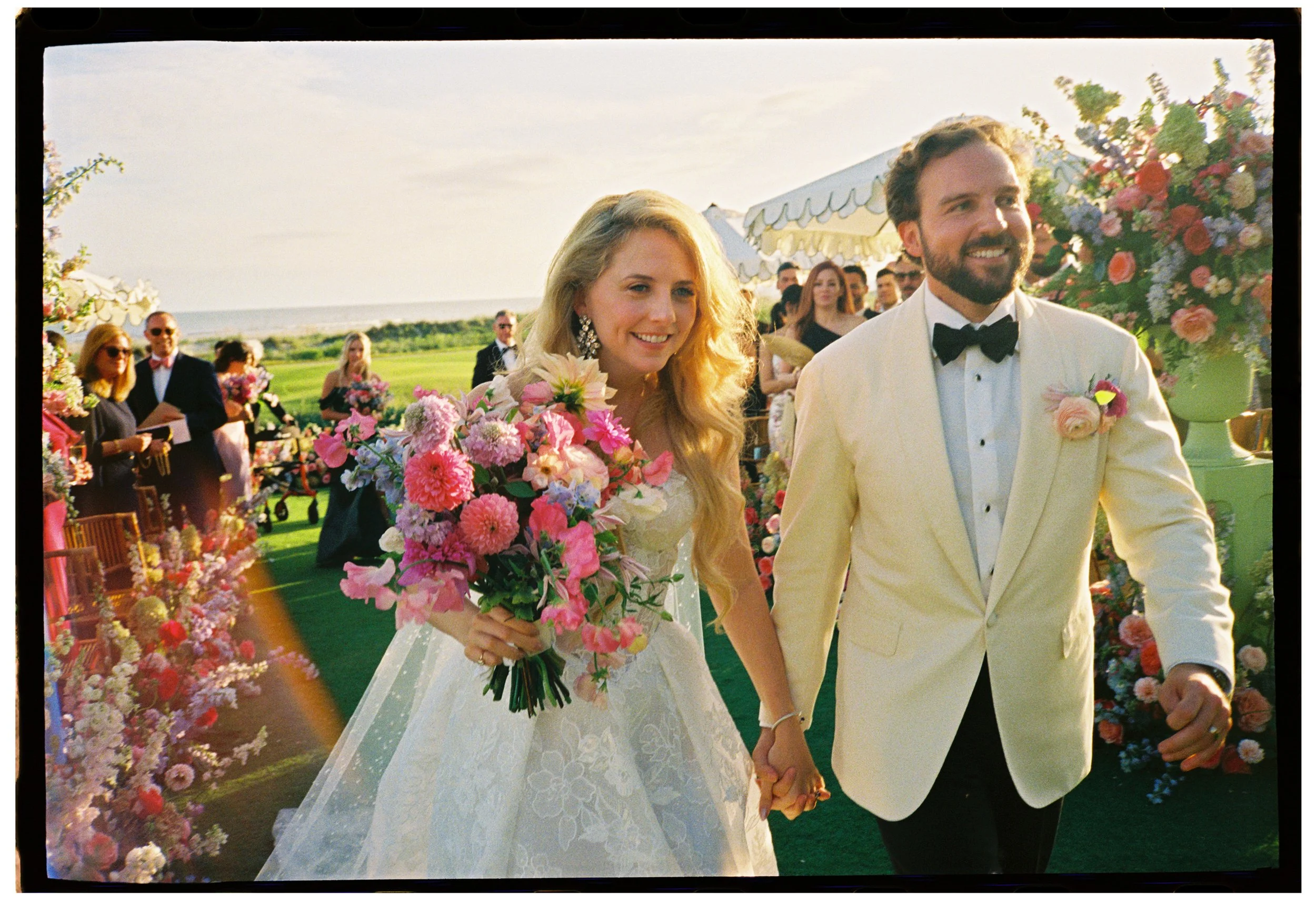 Film photograph of Katie and Trevor smiling and holding hands as they walk through their flower-filled recessional at The Ocean Course.