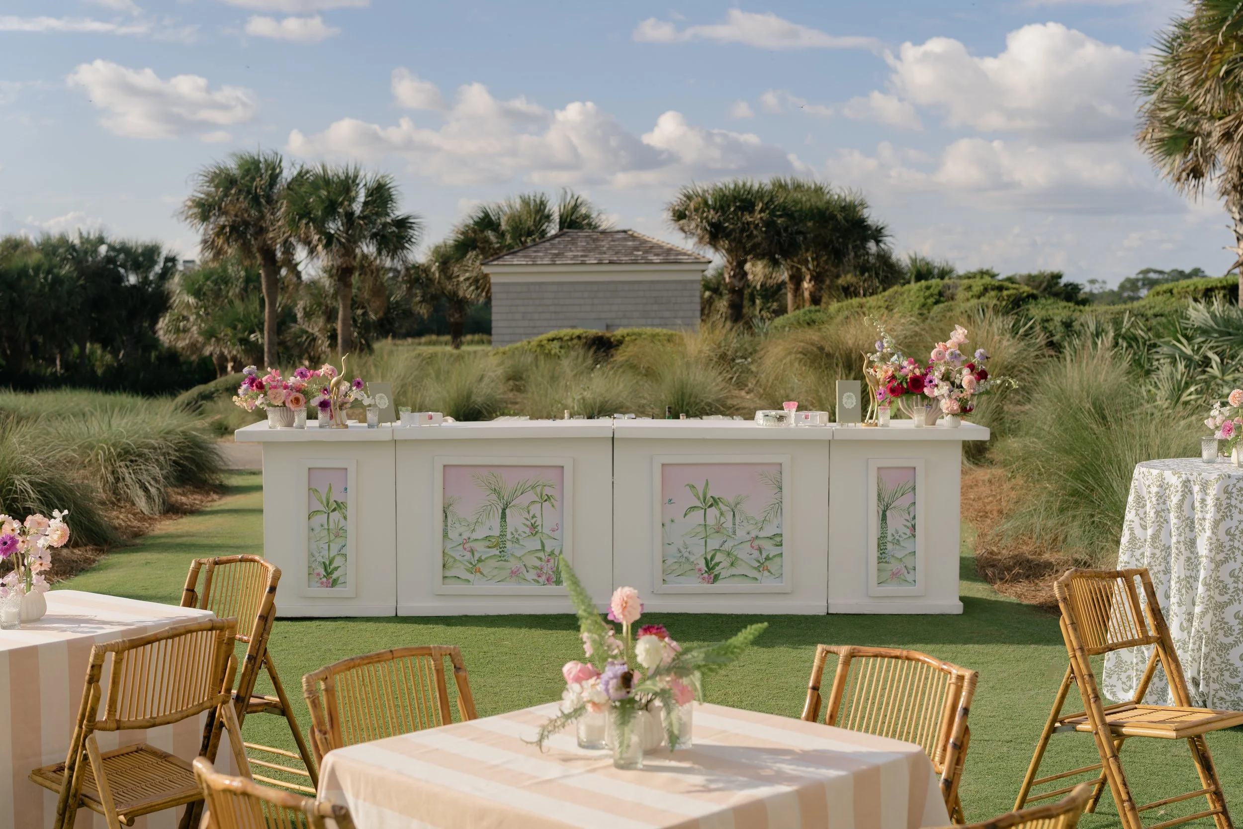 Custom white cocktail bar with tropical illustrated panels, pastel flowers, and rattan seating on the lawn at The Ocean Course.