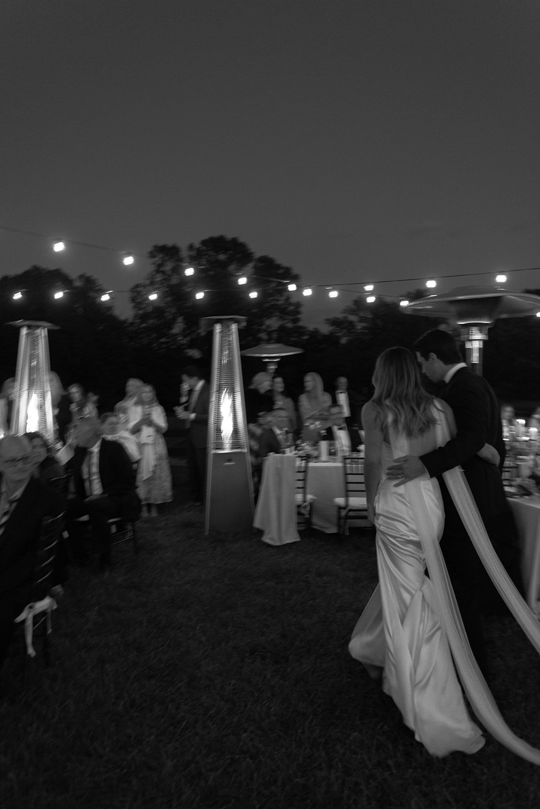 Black-and-white motion blur moment of the bride and groom walking through their outdoor ceremony at night beneath the string lights