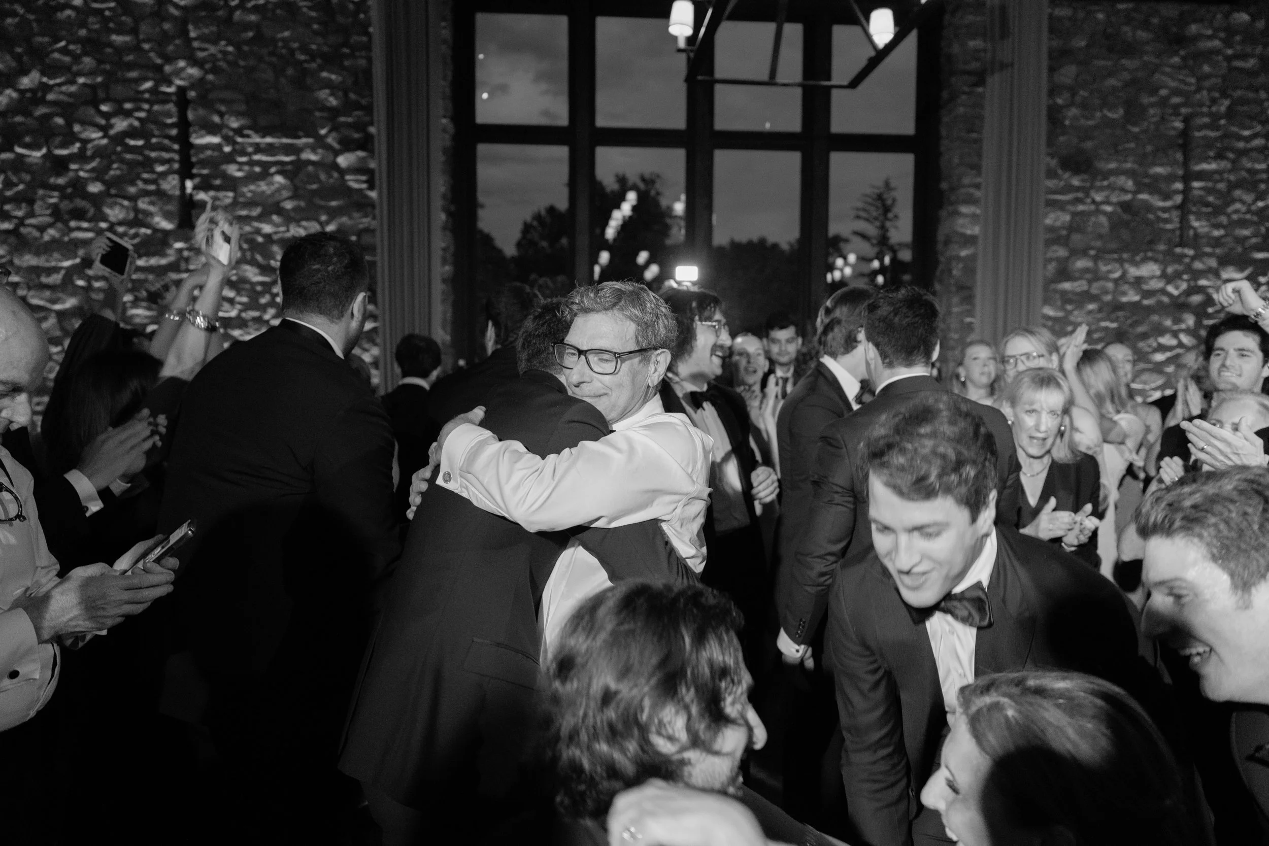 Black-and-white photograph of the groom and his father embracing on the packed reception dance floor.