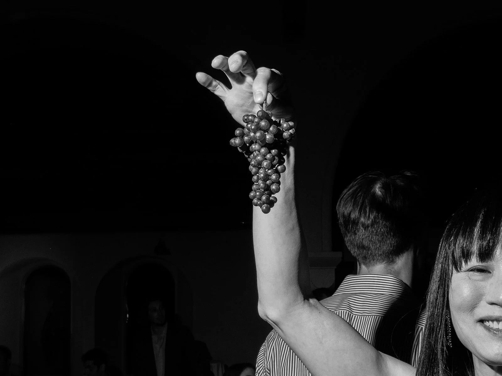 Black and white event photograph of a guest raising a bunch of black grapes on the vine during an evening celebration for a commercial event.