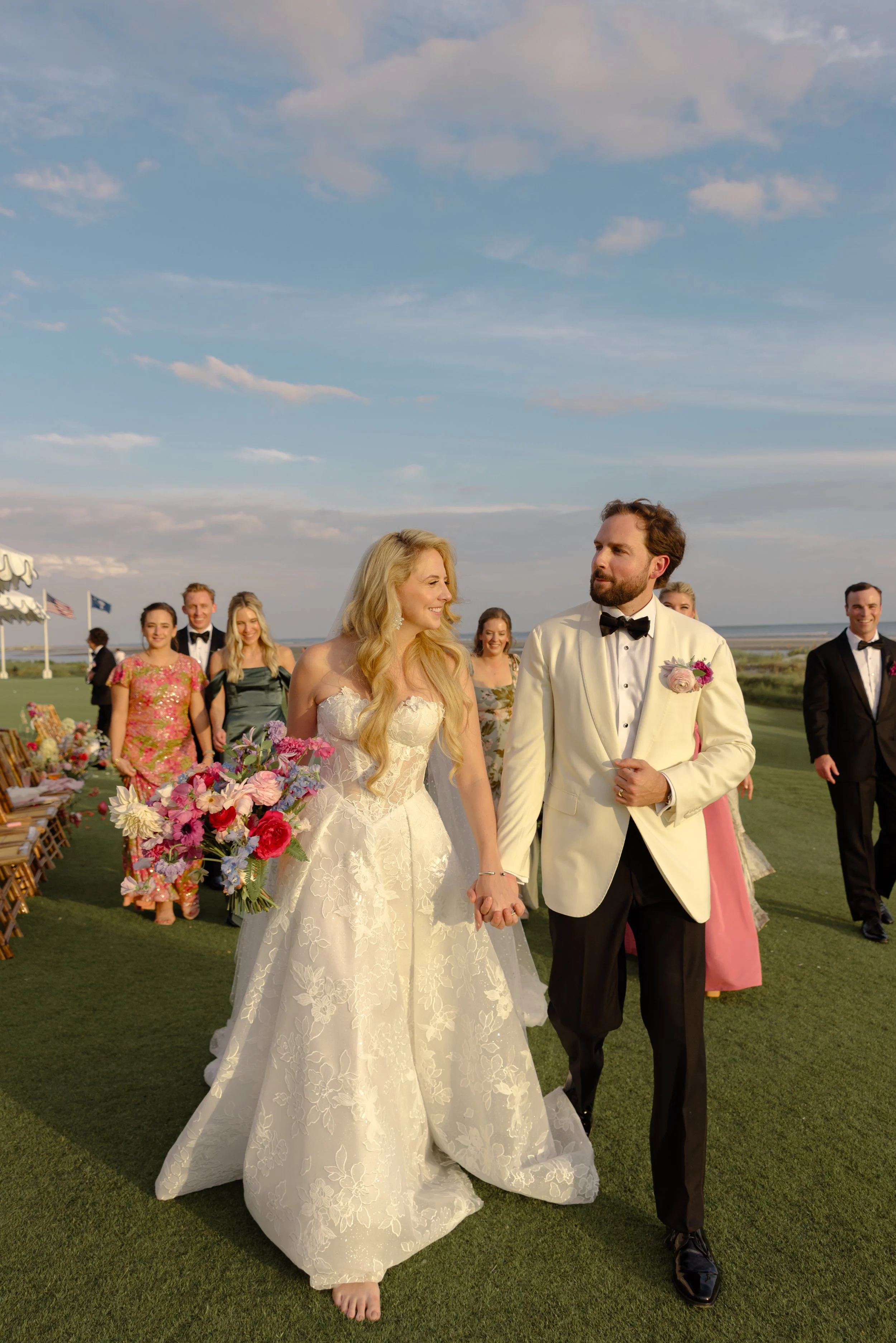 The bride and groom walk together across the Ocean Course lawn after their ceremony, surrounded by guests and colorful flowers.